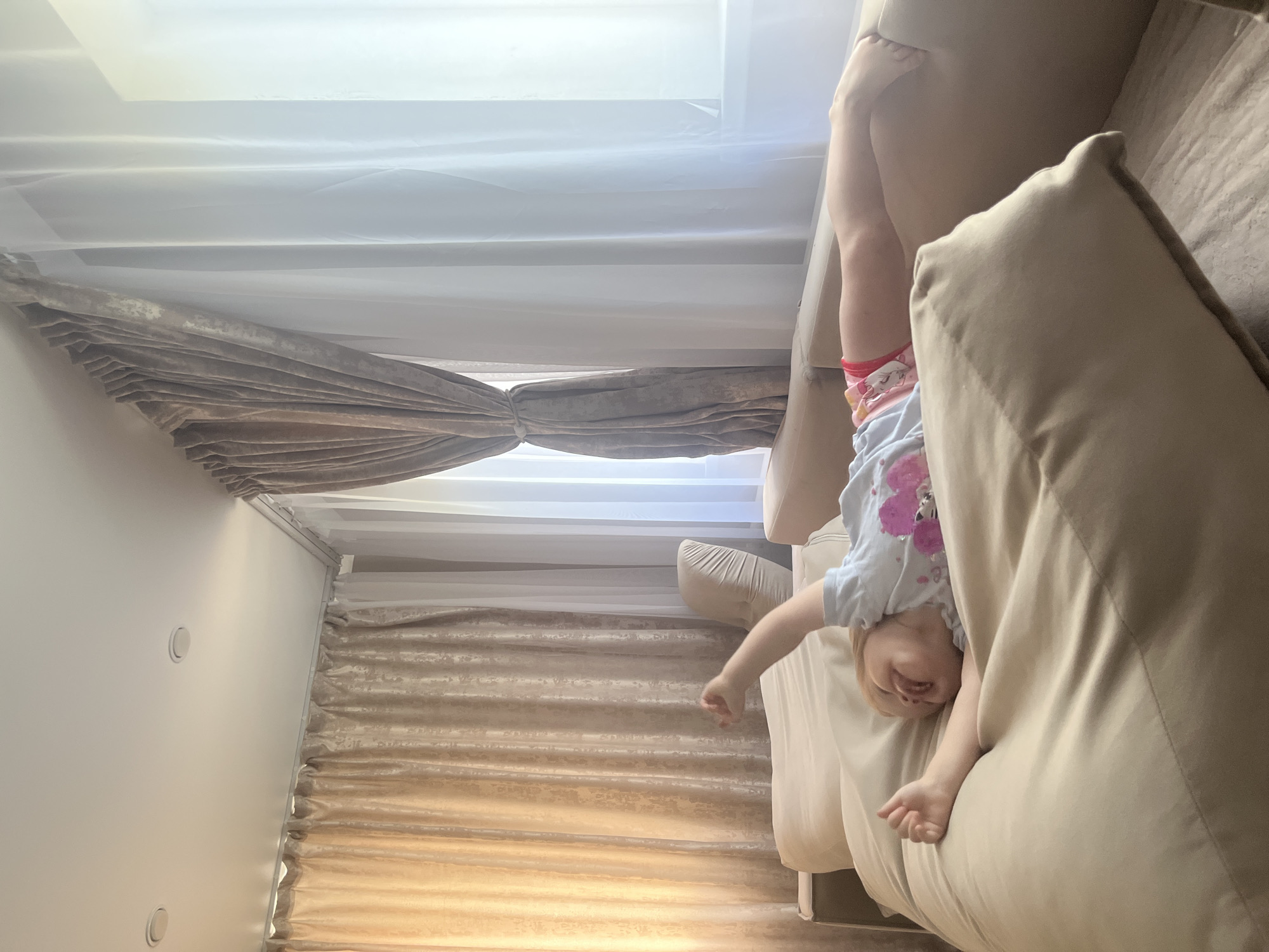 A young child with blonde hair is lying on a large, beige sofa, stretching their arms and legs. The child is wearing a light blue t-shirt with pink hearts and colorful shorts. The sofa is positioned in a room with large windows covered by beige and white curtains. The environment appears to be a residential interior, possibly a living room. The scene is brightly lit by natural light filtering through the curtains. There are no other people or animals visible in the image. The overall impression is one of a relaxed or playful moment. The location in Rakhinka, Russia, is not visually indicated in the image.