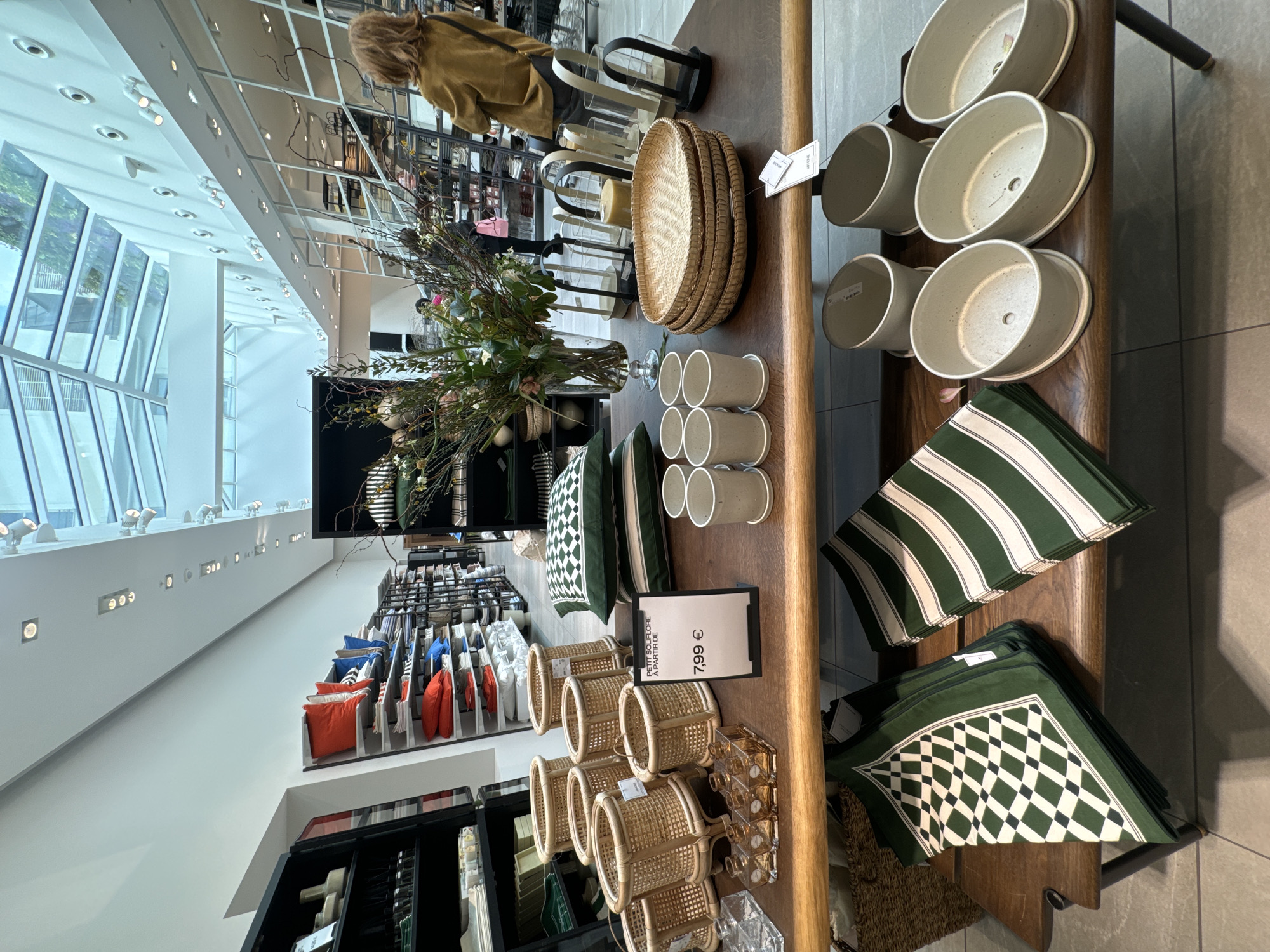 The image shows a display table in a store, likely in Paris, France. The table is made of wood and features various home decor items. There are several stacks of white ceramic pots, woven baskets of different sizes, green cushions with striped and checkered patterns, and glass candle holders. A price tag is visible on the table, indicating the cost of some items as "7,99 €." In the background, there is a flower arrangement in a vase, clothing racks with colorful textiles, and a person wearing a tan jacket. The store has large windows that allow natural light to flood the space. The overall impression is that the scene is a curated display of home goods in a retail environment.