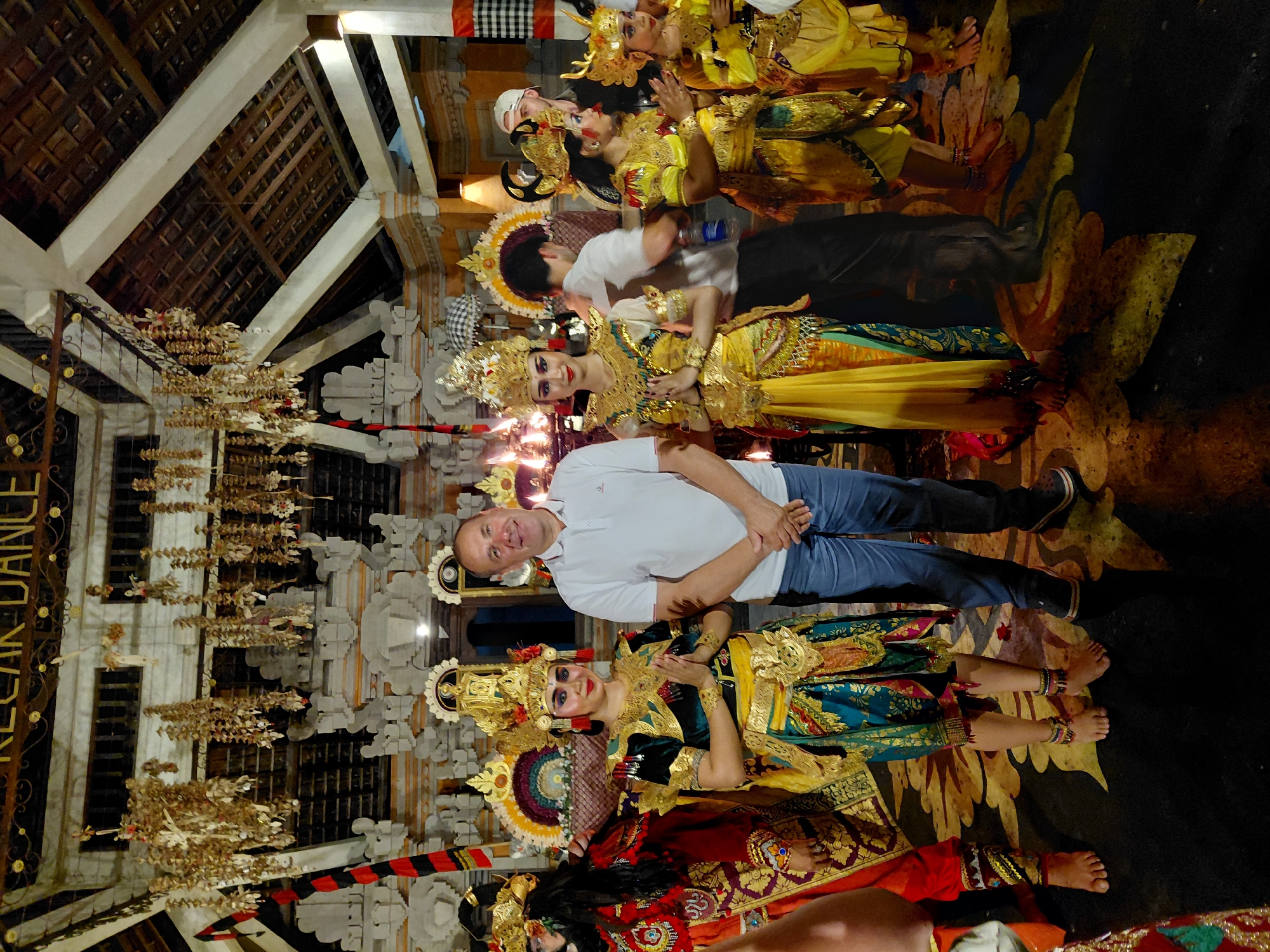 This media file depicts a group of people posing for a photo in what appears to be a cultural performance setting in Ubud, Indonesia. The subjects include several performers in elaborate traditional Balinese attire, characterized by ornate golden headdresses, vibrant yellow and green costumes, and detailed jewelry. They are positioned in a way that suggests they are either in the midst of or have just concluded a performance. A man in a white polo shirt and blue jeans stands among them, looking directly at the camera with a smile, seemingly a participant or observer interacting with the performers.
The scene is set indoors, likely a temple or a dedicated performance space, with visible architectural elements such as a thatched roof, intricate wooden beams, and decorative stone carvings. Hanging from the ceiling are traditional Balinese offerings or decorations made of dried leaves and flowers. The floor is dark and appears to be adorned with a large, stylized floral or mythical creature motif, possibly painted or inlaid. The lighting suggests it is nighttime or an indoor setting with artificial illumination, highlighting the colorful costumes and the architectural details. The overall mood is celebratory and cultural, with the performers displaying graceful poses and expressions.
Visible text in the image includes "KECAK DANCE" written vertically on a dark surface to the left of the frame, indicating the type of performance.