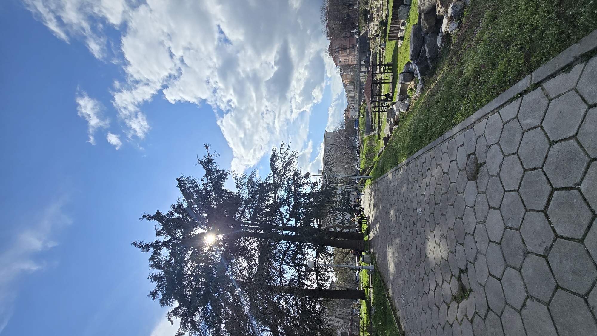 The image depicts a park scene in Stara Zagora, Bulgaria, with a paved walkway composed of hexagonal concrete blocks extending from the bottom right corner towards the center of the frame. Along the left side of the walkway are tall, mature trees with dense foliage partially obscuring the sun, creating a starburst effect. Beyond the trees is a clear blue sky with scattered white clouds. To the right of the walkway is a grassy area, interspersed with large rocks and picnic tables, before rising to meet the buildings of the city. A few people are scattered along the walkway, some sitting on benches and others walking. Several cars are parked along the street beyond the walkway. The lighting suggests it is a bright, sunny day.