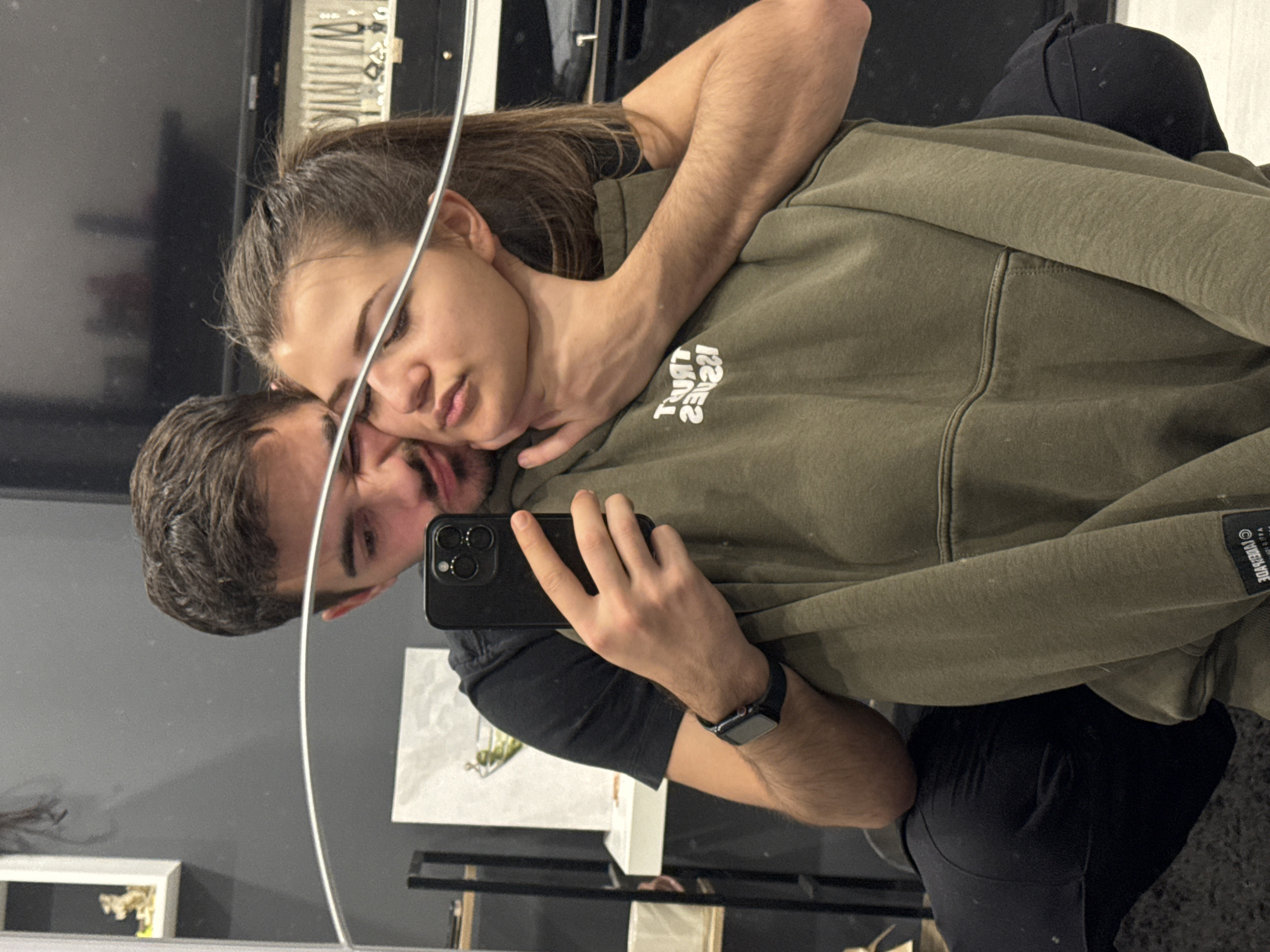 A couple is taking a selfie in a mirror. The man, with short brown hair, holds a black smartphone to take the photo. He wears a black T-shirt and a smartwatch on his left wrist. His face is partially visible in the mirror. The woman has brown hair pulled back in a ponytail and wears an olive green hoodie with white text. She leans her head against the man’s. The location appears to be a shop, with items on display behind the couple and below them in the mirror’s reflection. The area appears to be a retail space. The city is not identifiable.