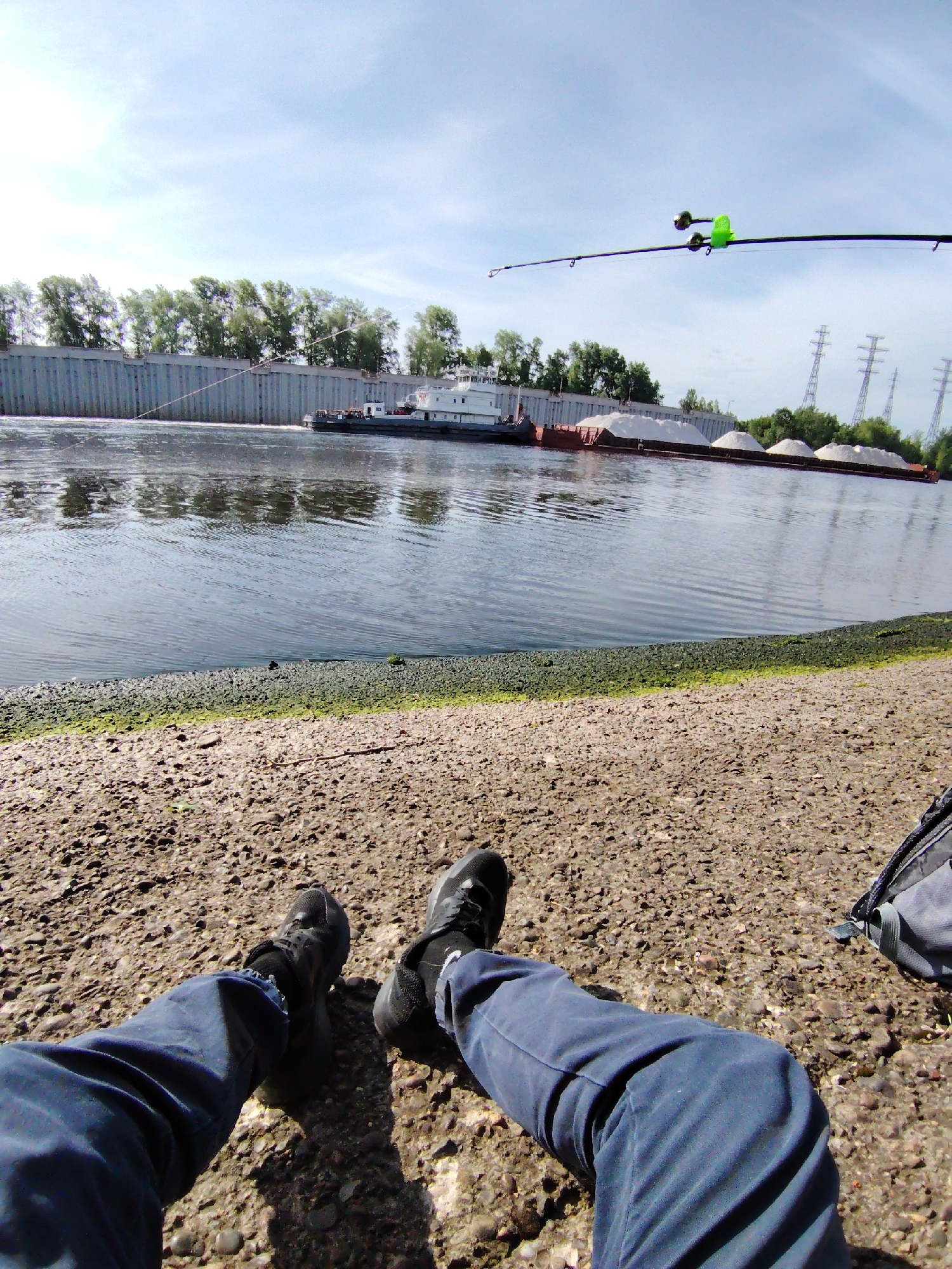 A first-person perspective photograph shows a person sitting on a gravelly shore in Chaikovsky, Russia, with a view of the Kama River. The individual is wearing blue pants and black shoes. A fishing rod extends into the upper right corner of the frame, suggesting the person is fishing. In the background, a tugboat pulls a barge loaded with what appears to be white aggregate (sand or gravel) along the river. On the opposite bank, a concrete wall and line of trees are visible, along with power line towers in the distance. The sky is partly cloudy.