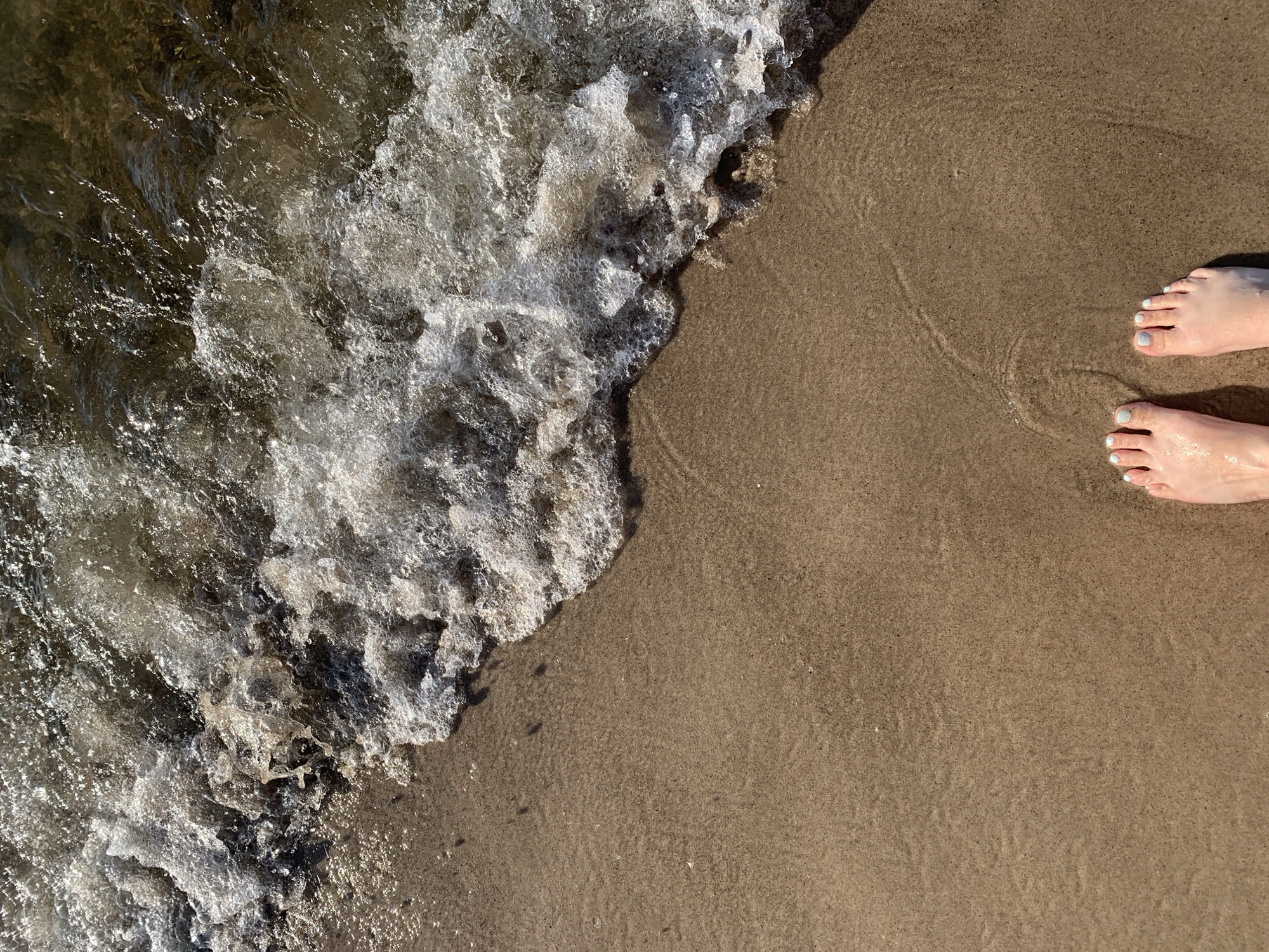 The image shows a close-up, top-down view of a sandy beach at the Baltic Sea coast, likely in or near Kołczewo, Poland. Two human feet, with pale skin and light blue nail polish, are positioned in the lower center of the frame, standing on damp sand. The sand exhibits subtle ripples and patterns from the receding water. The upper portion of the image is dominated by the edge of a wave, characterized by frothy white foam and clear, rippling water. The water reflects sunlight, creating bright, shimmering highlights. The overall scene depicts a serene moment at the seaside.