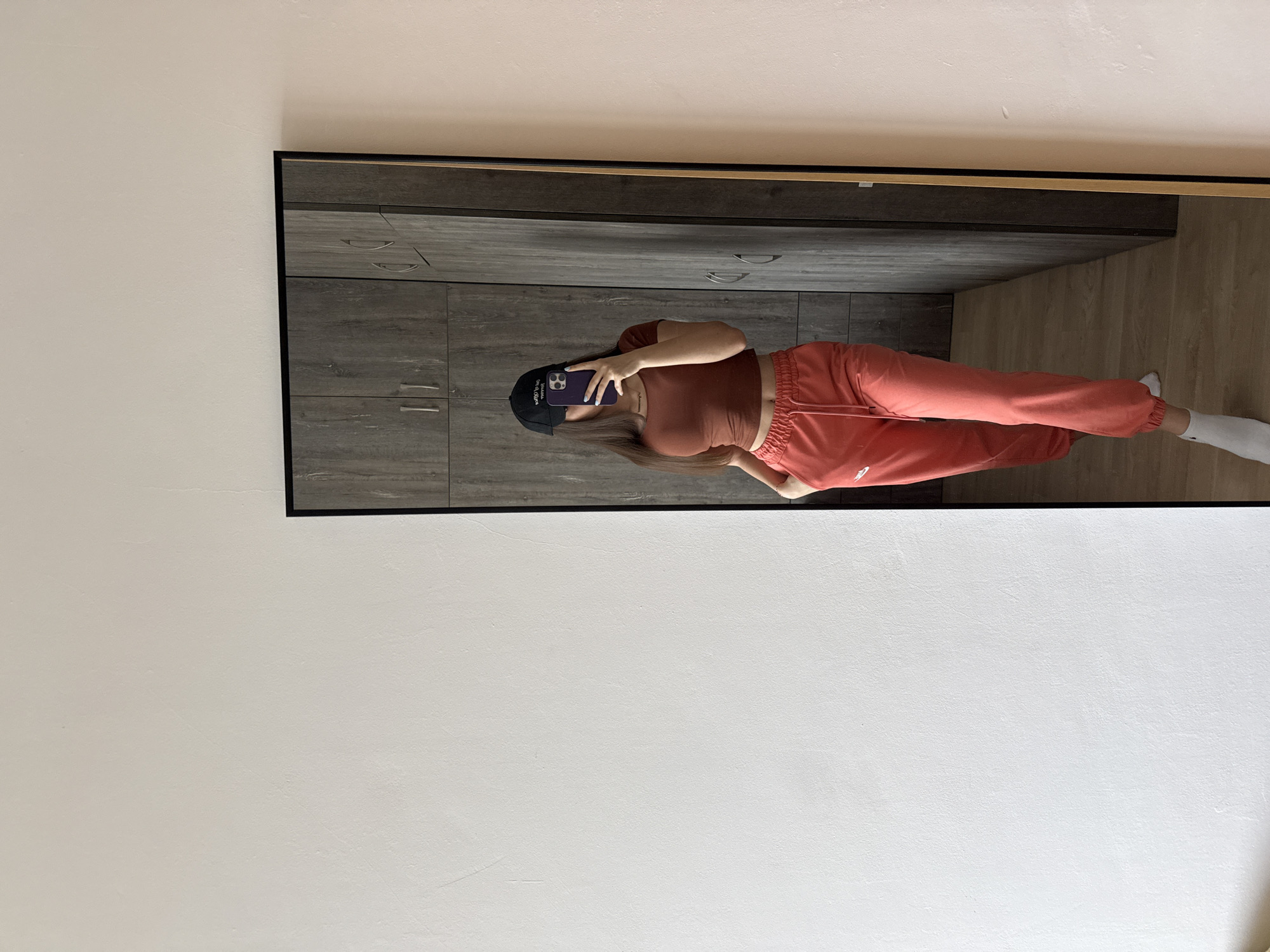 The image shows a person reflected in a mirror. The person is wearing a dark-colored baseball cap, a brown top, and reddish-orange sweatpants with white socks. They are holding a phone, presumably taking the photo. The background reflected in the mirror shows a gray cabinet and a light-colored wooden floor. The wall behind the mirror is white. The location is likely indoors. There is no way to tell if the location is in the city of Plovdiv, Bulgaria.
