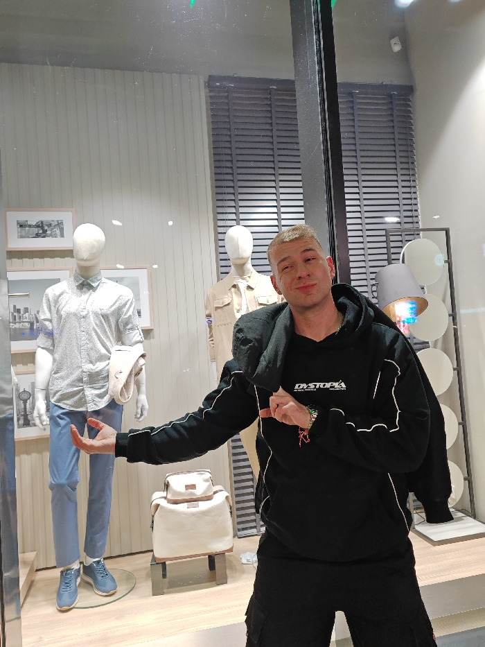 The image displays a male subject positioned in front of a retail clothing storefront display in Shumen, Bulgaria.
The subject is a young adult male with short, light-colored hair, facing the camera with a slight smirk and partially closed eyes. He is wearing a black hooded sweatshirt with white piping along the sleeves and sides, featuring the word "DYSTOPIA" printed in white on the chest. The hood appears to be resting on his shoulders. He wears dark cargo shorts or trousers. Multiple colorful bracelets are visible on his left wrist. His right arm is bent, with the index finger pointing towards his chest. His left arm is extended horizontally to the left, with the palm open and facing upwards.
Behind the subject, a glass window shows reflections and reveals the interior of a clothing store display. Three white, headless mannequins are visible inside. The leftmost mannequin wears a light-colored, patterned long-sleeved shirt with rolled-up sleeves, light blue trousers, and light blue sneakers; a cream-colored garment is draped over its left arm. A cream-colored backpack with brown accents rests on a dark display stand between the first two mannequins. The central mannequin, partially obscured, wears a tan collared shirt. The third mannequin is mostly obscured. The display features a vertically paneled background wall with several framed black-and-white urban landscape photographs. To the right, dark horizontal blinds are visible behind a window frame within the display, and a modern lamp with a gray shade and multiple circular, reflective elements stands as part of the decor. The flooring inside the display is light-colored.