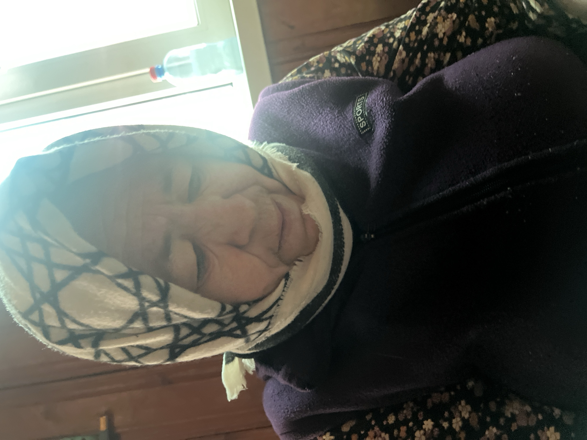 An elderly woman is seated indoors. She wears a white headscarf with a black geometric pattern, a dark purple fleece jacket with the word "SPORTS" on a tag, and a dark floral-patterned garment beneath. A window is visible in the upper left corner with a plastic water bottle on the sill. The location is Yuzhnoye, Russia.