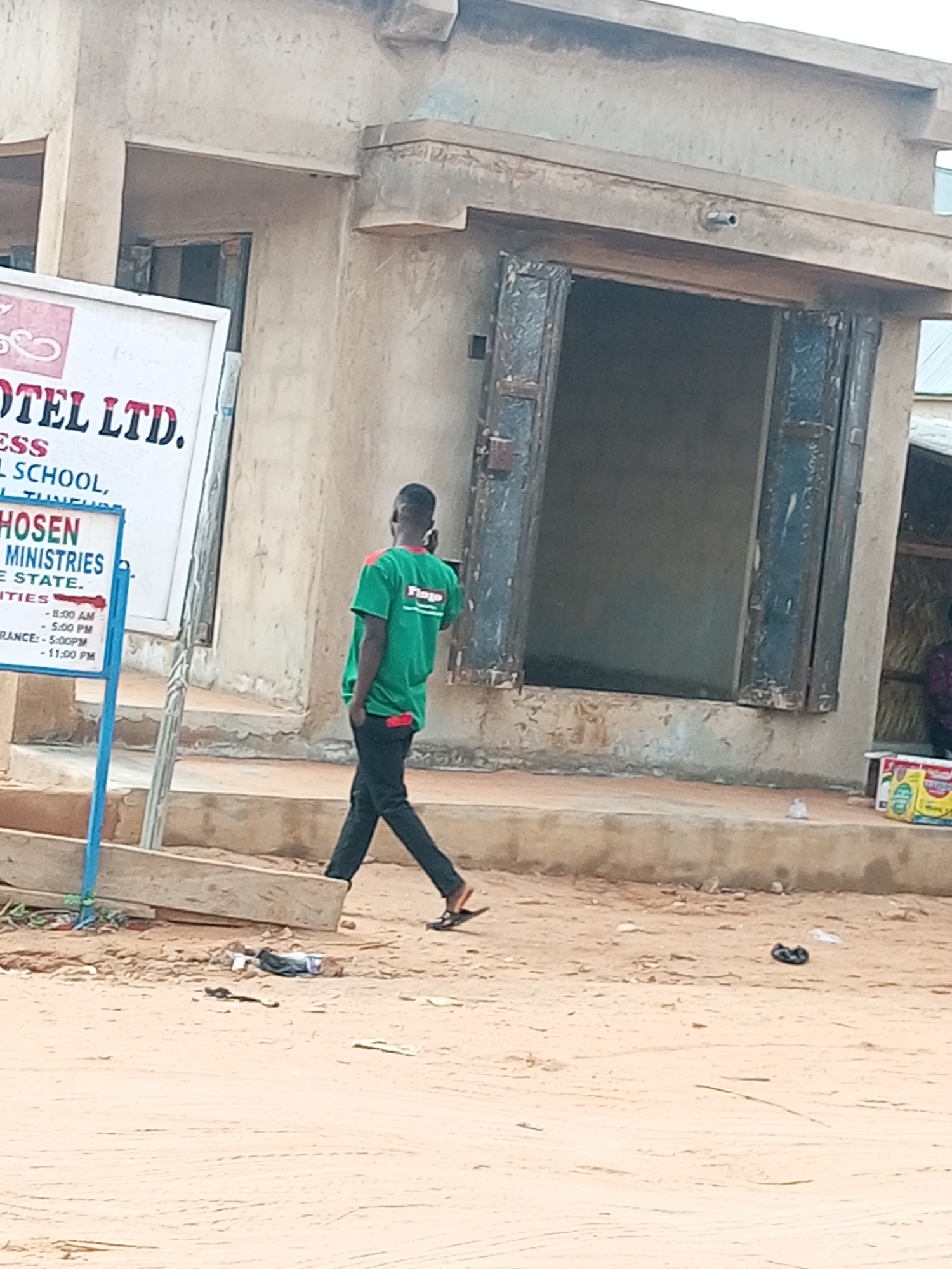 This media file features a man in a green shirt with "Fingo" printed on it, walking on a dirt path in front of a dilapidated building. The building appears to be made of concrete with a weathered facade and open, shuttered doorways. To the left of the man, a white sign with red and blue lettering is partially visible, displaying text that includes "OTEL LTD.", "SCHOOL", "MINISTRIES", and times such as "11:00 AM" and "5:00 PM". The ground is sandy and uneven, with some scattered debris. The scene suggests an outdoor setting in a rural or semi-urban area, possibly in Nigeria given the location context. The lighting indicates it is daytime, but the weather is not discernible. The man is walking away from the camera, and his posture is casual. There are no obvious signs of distress or urgency, suggesting a routine activity. The overall impression is one of a simple, everyday scene in a somewhat weathered environment.
