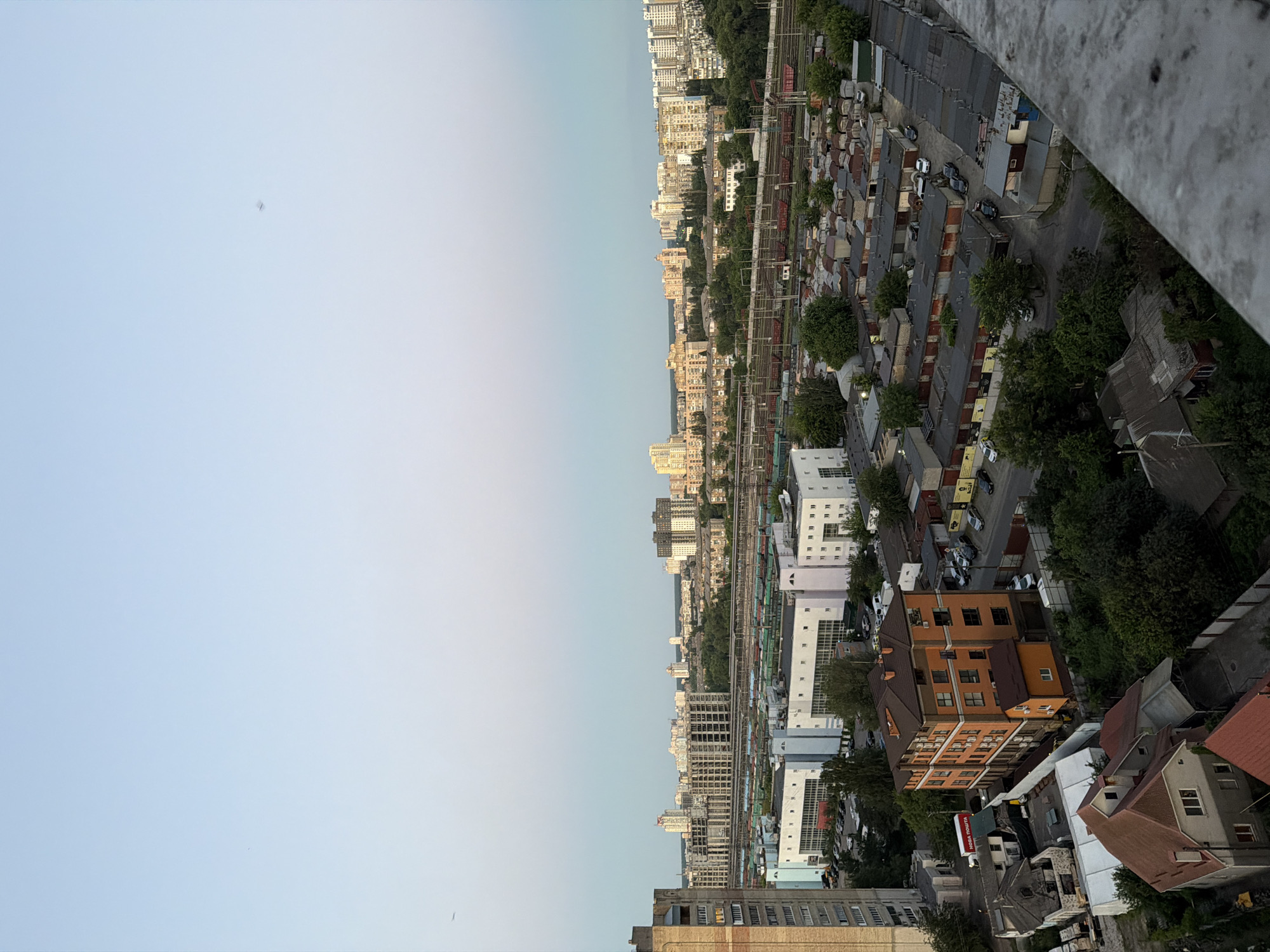 This is an aerial, high-angle, full shot of a city district in Kyiv, Ukraine, at dusk. The sky is a pale blue with a gradient to a lighter shade near the horizon. In the sky, there are two very small, faint birds or insects. The city landscape is dominated by a mixture of residential buildings, industrial structures, and a train track.
In the foreground, the edge of a concrete balcony or rooftop is visible, suggesting the viewpoint is from a tall building. Below, a cluster of small, weathered garages with red roofs stretches across the midground. They are arranged in rows on a paved surface, with several parked cars visible. Trees are interspersed among the garages.
Further into the midground, there is a long, multi-story building with white walls and numerous windows, likely an industrial or commercial facility. Adjacent to this building is a train track with several freight cars.
Beyond the industrial area, a dense urban panorama unfolds. Numerous apartment buildings of varying heights and architectural styles fill the background, with some under construction. The buildings are painted in shades of white, beige, and orange. Green trees are visible throughout the city, creating a contrast with the urban structures. A tall, brick apartment building is prominent on the far left side of the image. On the right side of the frame, nearer the foreground and between the garages and the apartment blocks, is an orange, multi-story residential building with a brown roof. Adjacent to it is a smaller orange structure and some smaller houses with red roofs.
The overall impression is of a developed urban area with a mix of residential, industrial, and transportation infrastructure, viewed from a height at the end of the day.