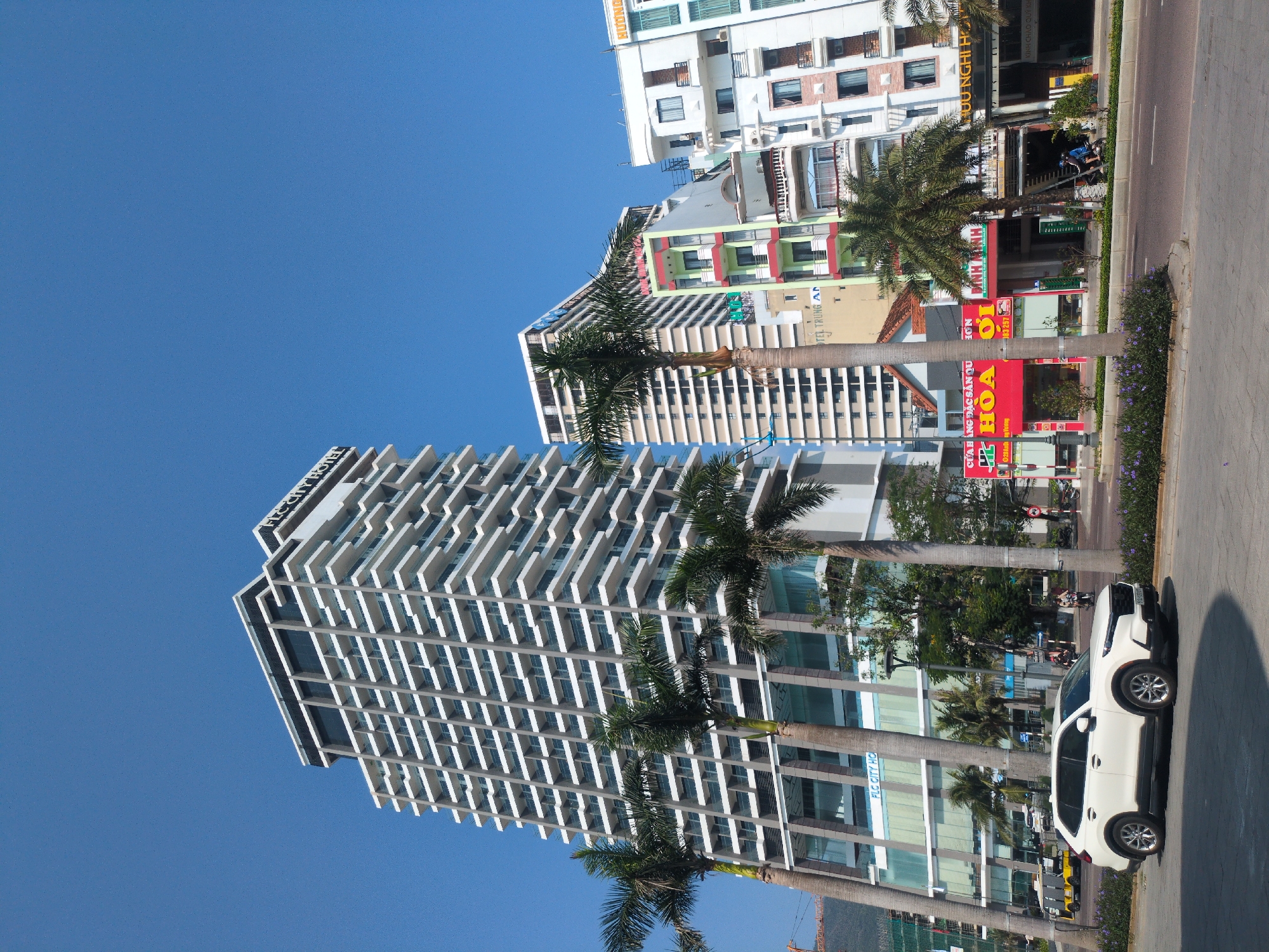 The image depicts a street scene in South Quy Nhon Ward, Vietnam. The frame is dominated by several high-rise buildings, including a modern, white tower with a unique balcony design. Palm trees line the street, obscuring partial views of the buildings. Street-level establishments include shops with brightly colored signage in Vietnamese, such as "HOA," "BÌNH MINH," and "HƯƠNG NGHỊ H." A white SUV is parked on the side of the road. Several people are traveling on motorbikes in the distance. The sky is clear and blue, suggesting a sunny day.