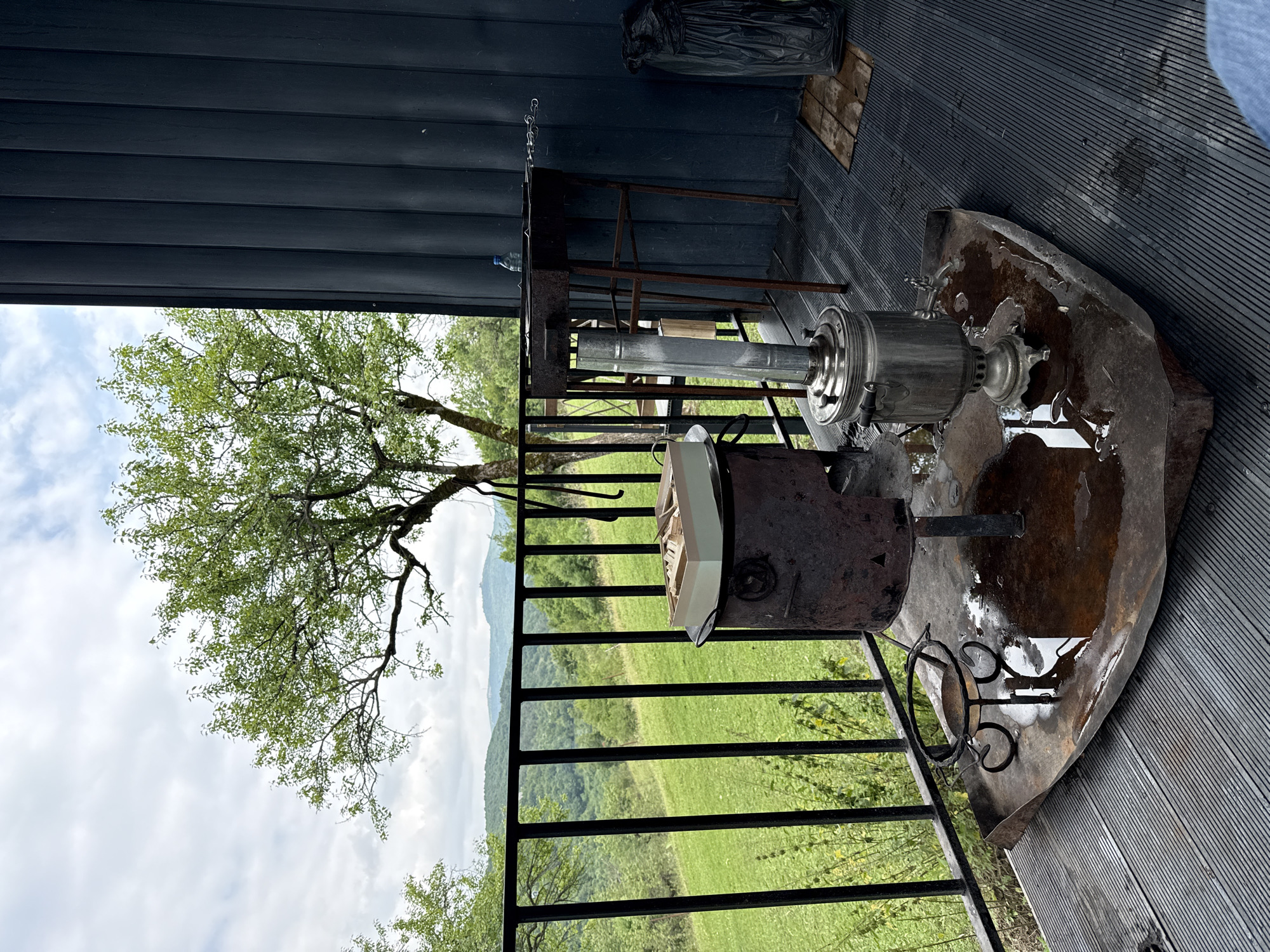The image depicts an outdoor setting, likely a balcony or porch, overlooking a lush green landscape with rolling hills and trees under a cloudy sky. In the foreground, there are several objects associated with outdoor cooking and hospitality. A large, rustic metal brazier sits on a weathered metal tray that has collected some water. On top of the brazier, there is a light-colored box containing what appear to be wooden skewers or utensils. To the right of the brazier is a traditional metal samovar, a device for heating and boiling water for tea, also situated on the metal tray. Behind the samovar and brazier, a rusty metal grill stand holds a grill grate and a single skewer. A dark-colored, ribbed wall, likely part of a building, forms the backdrop on the right side of the image. A black plastic bag is visible near the wall. The ground surface appears to be made of dark, slatted material. The scene is set in the city of Sis, Azerbaijan. No people are visible in the image.
