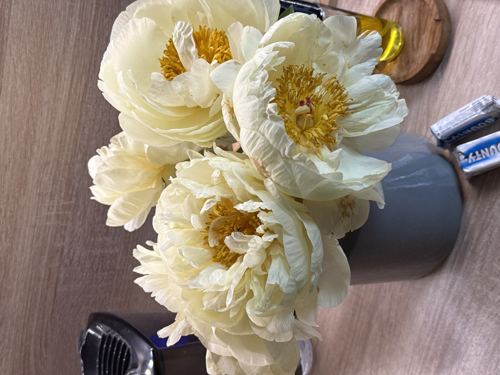 The image depicts a bouquet of white and yellow peonies in a gray vase, resting on a wooden surface. Next to the vase are two small Bounty chocolate bars. A black object with grill-like details is visible in the lower left corner. In the background, there's a wooden coaster and a small amber-colored glass object. The location isn't apparent from the visual data, so assuming the location is Krasnoyarsk, Russia is not possible.
