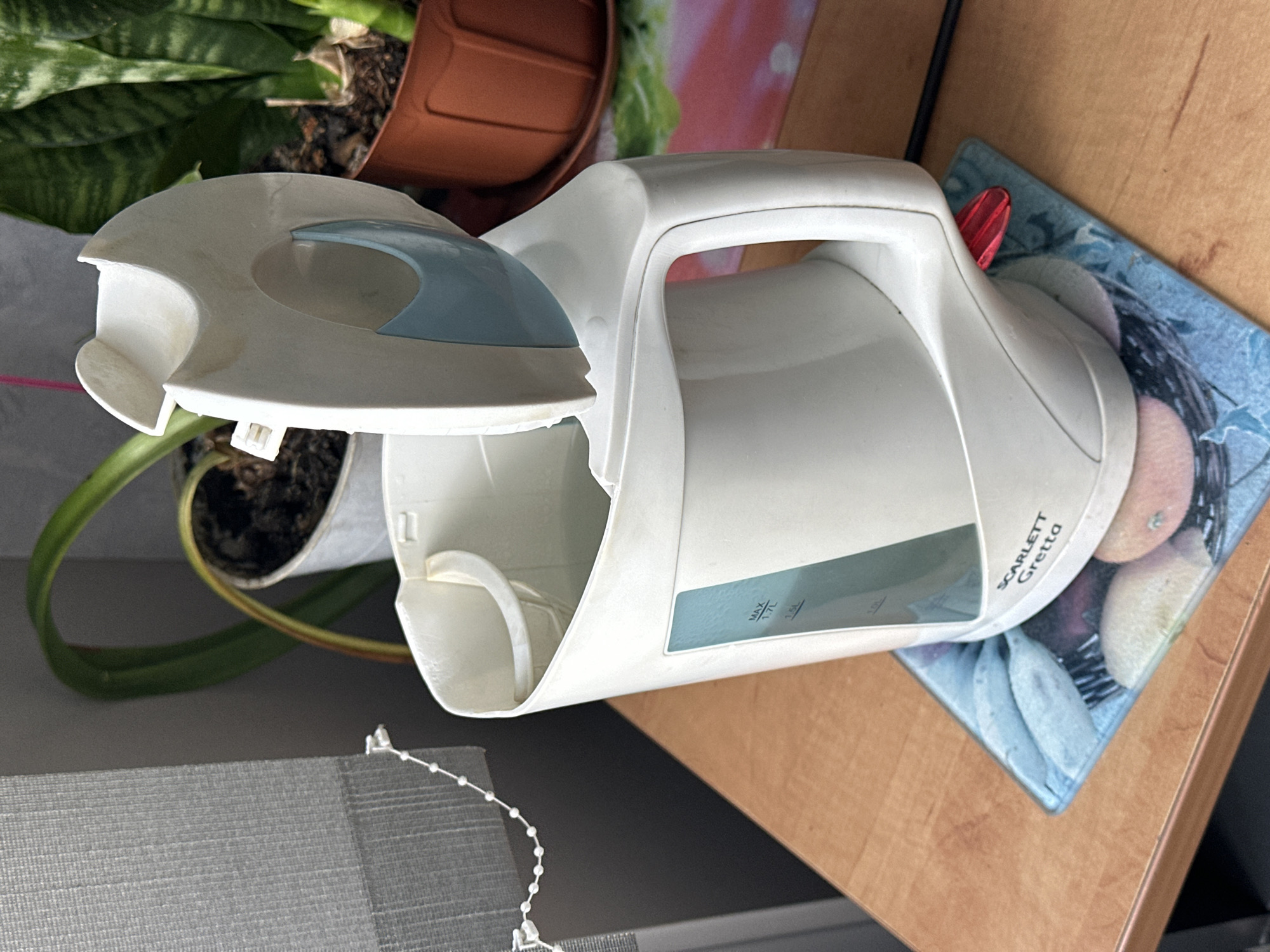 The image shows a white electric kettle with its lid open, sitting on a wooden table. The kettle is placed on a decorative mat with images of fruit. The kettle has a blue water level indicator with markings for "MAX 1.7L", "1.5L", and "1.0L". The brand name "SCARLETT Gretta" is visible on the kettle. In the background, there are potted plants, one of which appears to be a snake plant, and a grey roller blind. The scene is indoors, with natural light filtering in from a window. The context of Hora, Ukraine, is not directly evident from the visual elements within the image itself.