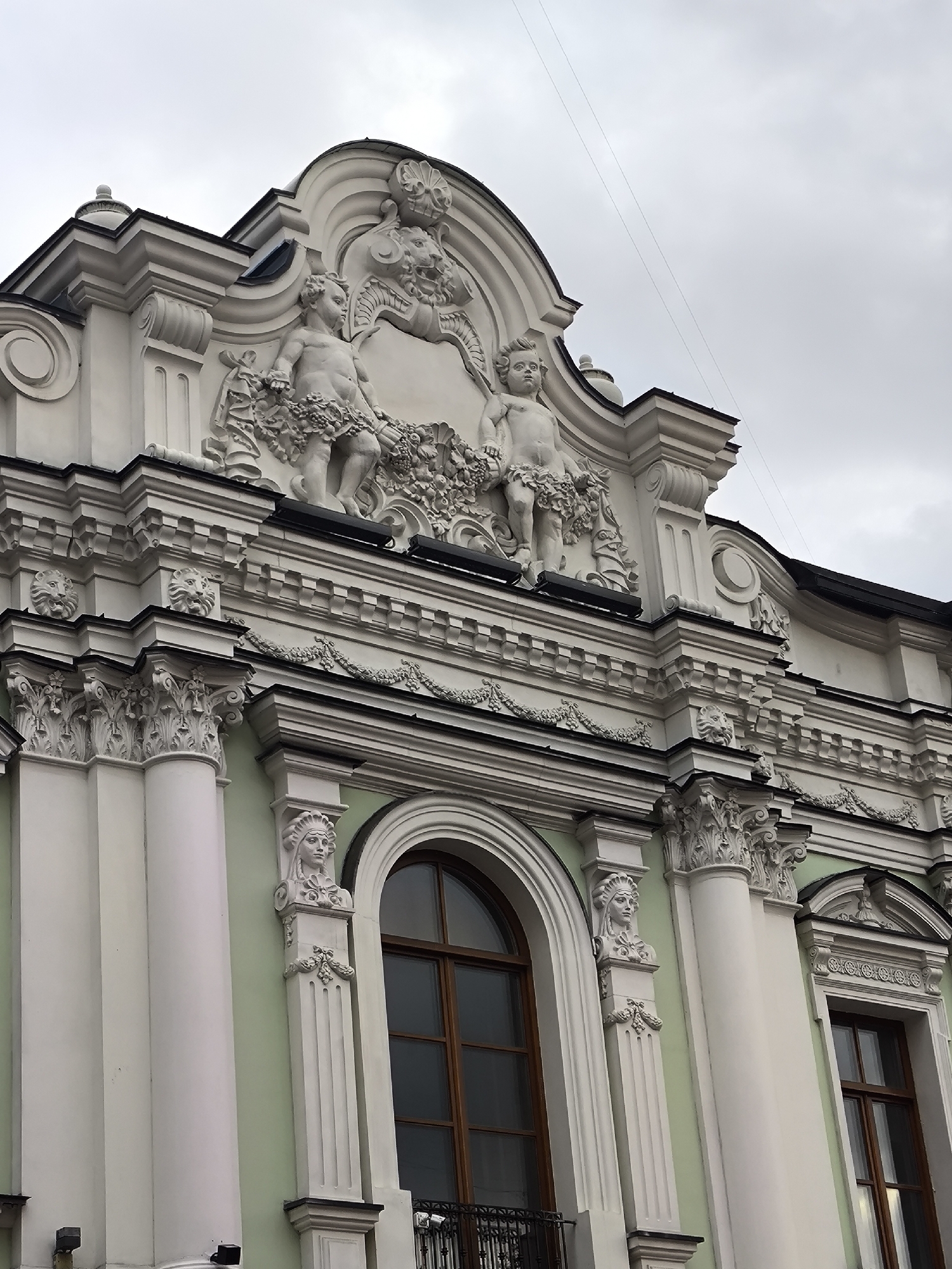 The image displays the ornate upper facade of a building in Moscow, Russia, under an overcast sky. The architecture features numerous white decorative elements contrasted against a light green wall.
At the highest visible point, a curvilinear pediment is adorned with high-relief sculptures. Centrally, two cherubic figures are depicted, each holding abundant garlands of flowers and fruits. Behind them, and integrated into the pediment's design, are additional floral motifs and a stylized lion's head relief visible on the right.
Below the pediment, a detailed frieze extends horizontally across the facade, showcasing repeating patterns and multiple smaller lion head reliefs. The building's structure is articulated by robust, fluted columns topped with elaborate Corinthian or composite capitals. Between these columns, tall arched windows with dark wooden frames are visible, and a small wrought-iron balcony railing can be seen beneath one window. Further decorative pilasters flank the windows, each surmounted by a sculpted bust of a female figure wearing a distinctive, ornate headpiece that suggests a stylized classical or indigenous influence.
No human activity is depicted in the static architectural scene. The sky is a uniform gray, indicating an overcast day. Thin electrical wires are faintly visible crossing the sky. There is no visible text on the building.