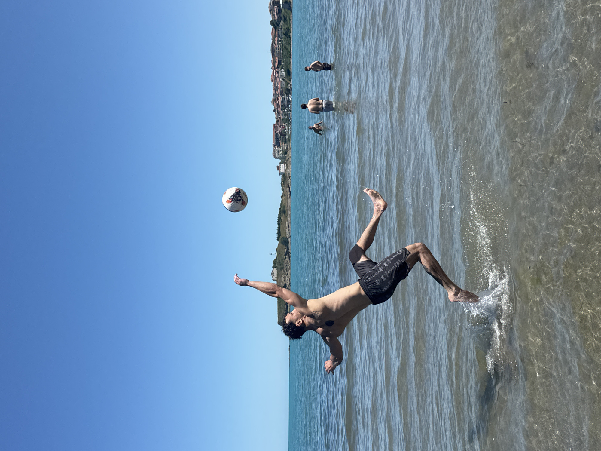 This media file depicts a sunny day at a beach in Tsarevo, Bulgaria. The scene is set on a clear blue sky day, with the sun glinting off the shallow, clear water. In the foreground, a young man is captured mid-air, performing a somersault in the water, with a soccer ball suspended beside him. He is wearing black swim shorts with a repeating pattern. His body is arched, and his arms are outstretched, suggesting an athletic maneuver. Water splashes around his feet as he is about to land or has just pushed off from the water's surface.
In the background, several other people are visible in the water, wading and enjoying the sea. They appear to be a distance away from the main subject, positioned further into the water. Beyond the water, a coastal town with buildings clustered on a hillside stretches along the horizon, identifiable as Tsarevo. The architecture suggests a Mediterranean or Eastern European coastal town. The overall atmosphere is one of leisure and recreation, with the bright sunlight and clear water indicating pleasant weather, likely during the summer months. The image captures a dynamic moment of sport and fun in a picturesque coastal setting.
