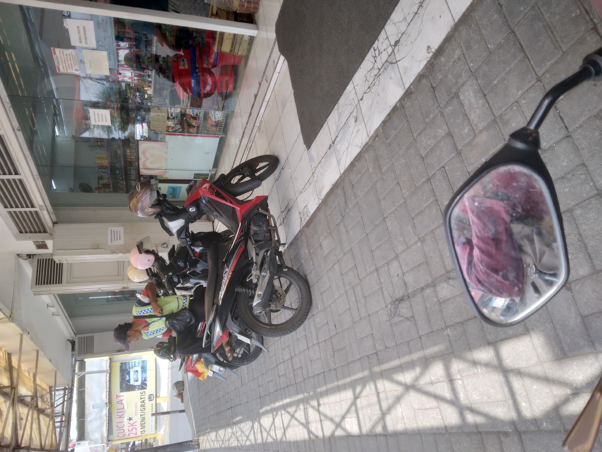 The image depicts a street scene in front of a shop in the city of Berkoh, Indonesia. The foreground is dominated by the reflection in a motorcycle's side mirror, showing a person wearing a red patterned shirt. The middle ground features two motorcycles parked on a paved sidewalk. The one on the right is a red and black model, and the one on the left is black with a yellow flag. A person wearing a yellow vest and a cap is seated on the black motorcycle. The background shows the storefront of a shop with large glass windows revealing shelves stocked with various products. A yellow sign with text in Indonesian is visible on the left side of the image, near a white car graphic. The ground is paved with interlocking grey bricks, and a dark mat is placed in front of the shop's entrance. The overall lighting suggests it is daytime.