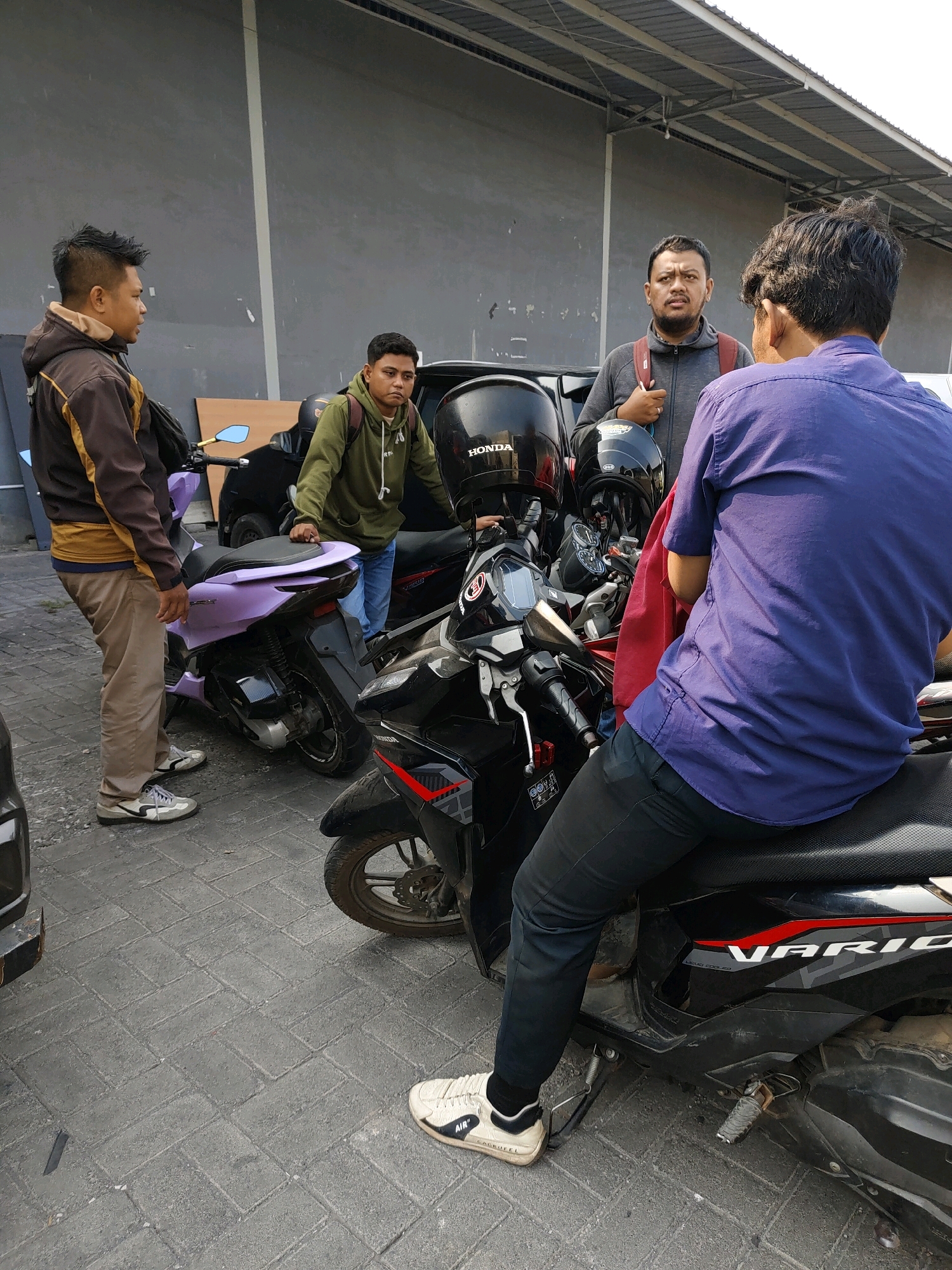 In Probolinggo, Indonesia, four men are gathered near several parked motorcycles. Two men are standing, one in a brown and yellow jacket and khaki pants, the other in a green hoodie and blue jeans, engaged in conversation. Another man, wearing a gray hoodie and a red backpack, stands between them, looking towards the camera. The fourth man, seen from the back and seated on a black motorcycle, wears a blue button-down shirt and dark pants. The environment appears to be an outdoor parking area, possibly adjacent to a building with a gray, textured wall and a corrugated metal roof overhead. The ground is paved with interlocking gray bricks. One motorcycle is light purple, while others are black, with helmets placed on them. The scene suggests a casual gathering or a moment before or after riding motorcycles.