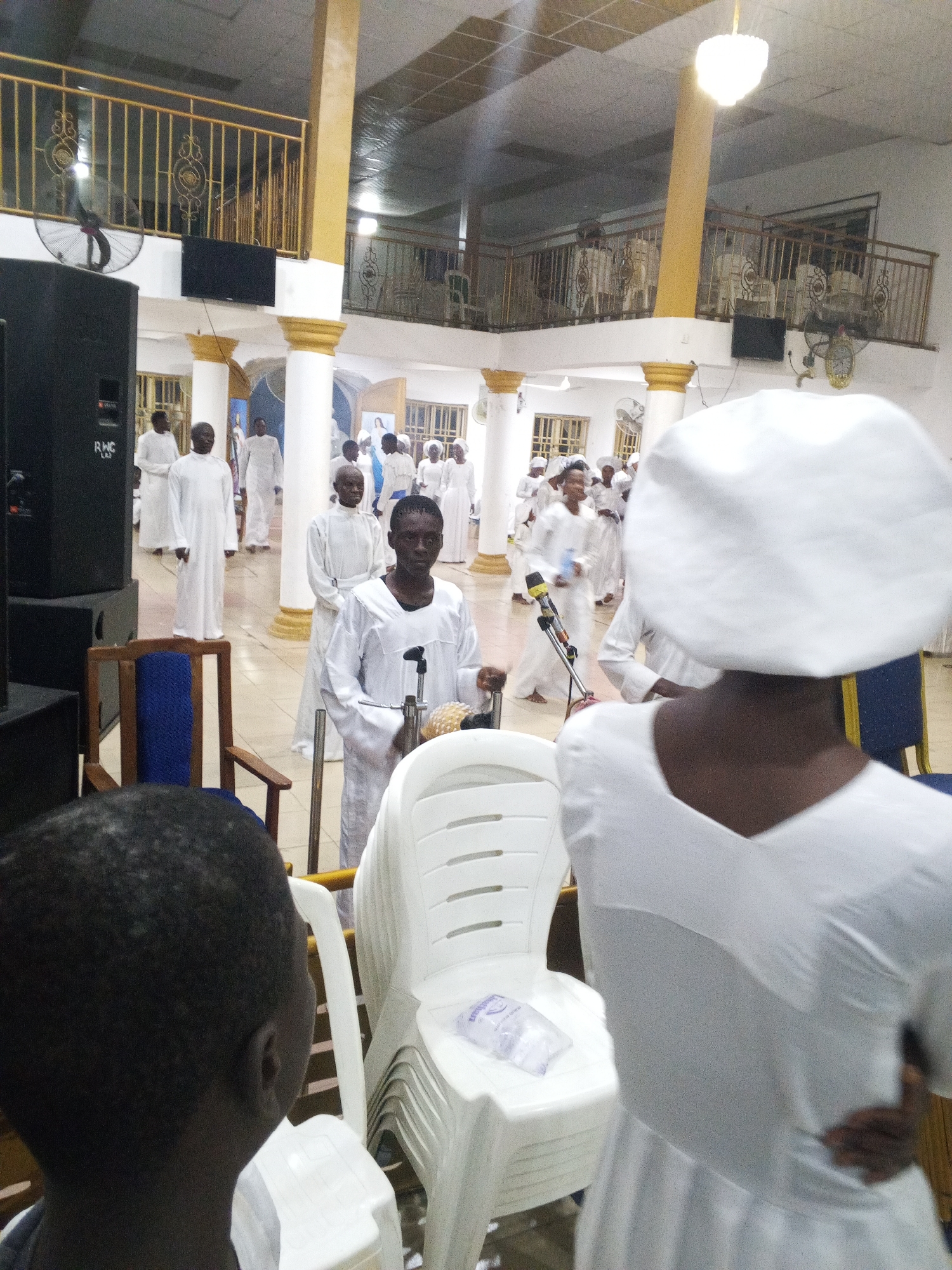 The image shows a gathering of people inside a building in Lokoja, Nigeria. The people are mostly dressed in white garments. Some are moving around, while others are standing still. The building appears to be a church or religious assembly hall with pillars and a balcony. There are chairs, sound equipment, and fans visible.