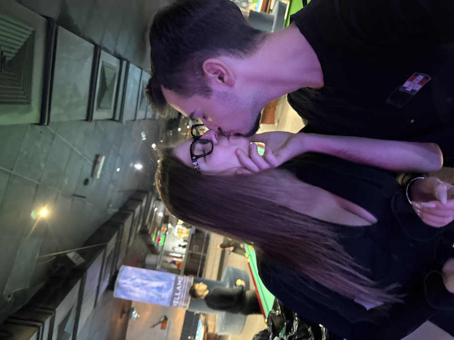 Two individuals, a man and a woman, are depicted kissing in the foreground of what appears to be an indoor entertainment venue. The woman, with long brown hair and wearing black-rimmed glasses, is dressed in a black, low-cut zippered top and holds a clear glass with a beverage in her left hand. Her right hand is placed on the man's chin/jaw area. The man, with dark hair and slight facial stubble, wears a black t-shirt featuring a "TOMMY" brand logo on the left chest. Both subjects are illuminated with a purple hue, suggesting specific ambient lighting.
In the background, multiple pool tables with green felt surfaces are visible, indicating a billiards establishment. A tall, illuminated vertical sign on the left background clearly displays the word "VELLAMO". Additional blurred figures are present throughout the background, engaged in various activities around the tables and what appears to be a bar area further back. The ceiling features an industrial design with visible rectangular ventilation grates and recessed lighting. Based on the "VELLAMO" signage, the location is identified as Vellamo Club or Vellamo Bowling & Billiards in Sofia, Bulgaria.
