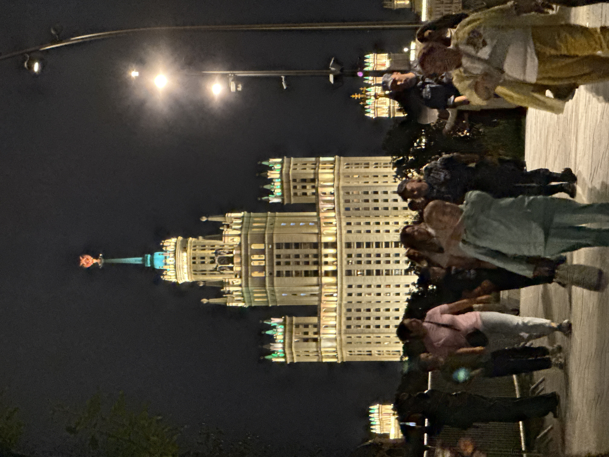 In Moscow, Russia, a large, illuminated Stalinist skyscraper dominates the night sky, its spire topped with a red star. The building is surrounded by smaller illuminated structures. A group of people are walking on a pedestrian walkway in the foreground, illuminated by streetlights. The people are of various ages and are dressed in casual clothing. Some are walking together, while others are walking alone. The overall atmosphere is one of urban nighttime activity.