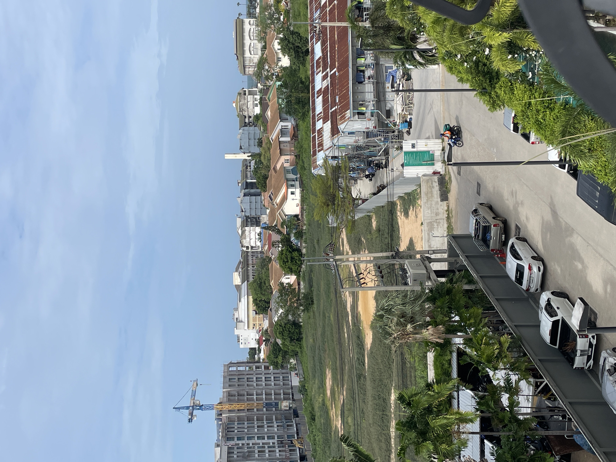 This is an eye-level, full shot of a street scene in Pattaya City, Thailand, with a view of buildings and a sky with scattered clouds. In the foreground, there are palm trees and a dark metal railing. To the right, a person rides a blue motorcycle with an orange box on the back. Further back, white cars and trucks are parked along the street, and a row of buildings with corrugated metal roofs is visible. In the middle ground, there is a large, empty field with some overgrown grass and dirt patches, bordered by trees. To the left, a tall, unfinished concrete building stands with a blue and yellow construction crane behind it. In the background, a cluster of houses with terracotta roofs and several taller buildings, including a white tower-like structure, are visible, suggesting a mix of residential and commercial areas. The scene appears to be a typical urban landscape with ongoing construction.