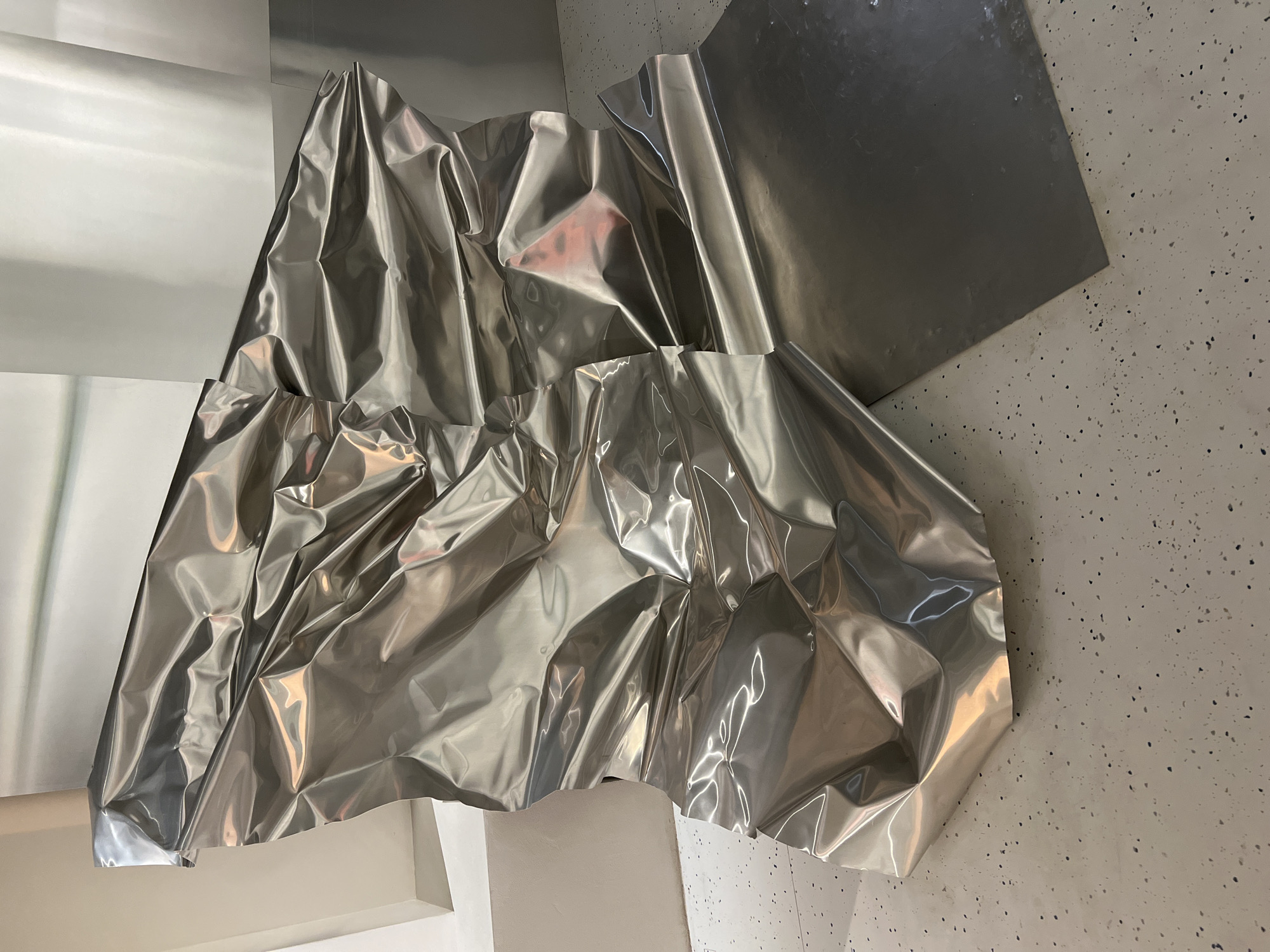 A photograph showcases an art installation in what appears to be a gallery space. The primary feature is a series of large, reflective, crumpled silver metallic sheets arranged to create an abstract, sculptural effect. One section appears to be shaped like a folded or draped fabric, while other sheets are flat and mounted on the wall. The backdrop is a textured wall with a speckled pattern and a smooth, light-colored surface. A part of the wall is angled with a corner. The location is Moscow, Russia.