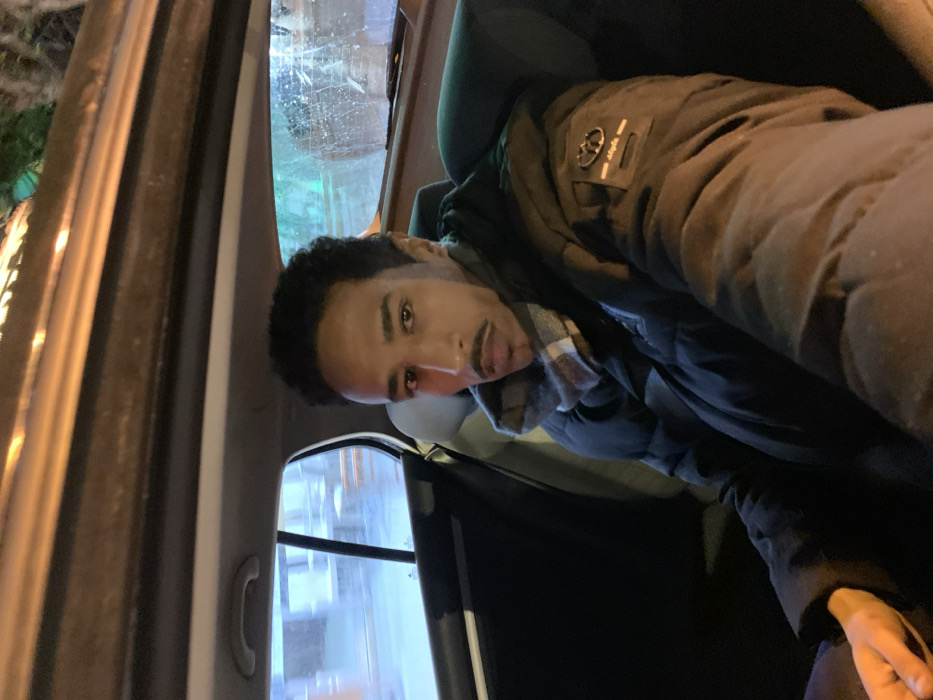 A man with short, dark hair and a mustache sits in the back of a car. He is wearing a dark, puffy jacket over a plaid shirt. The car's window and part of the interior are visible. The lighting is dim, suggesting it is nighttime or a dimly lit environment. The man's expression appears neutral.