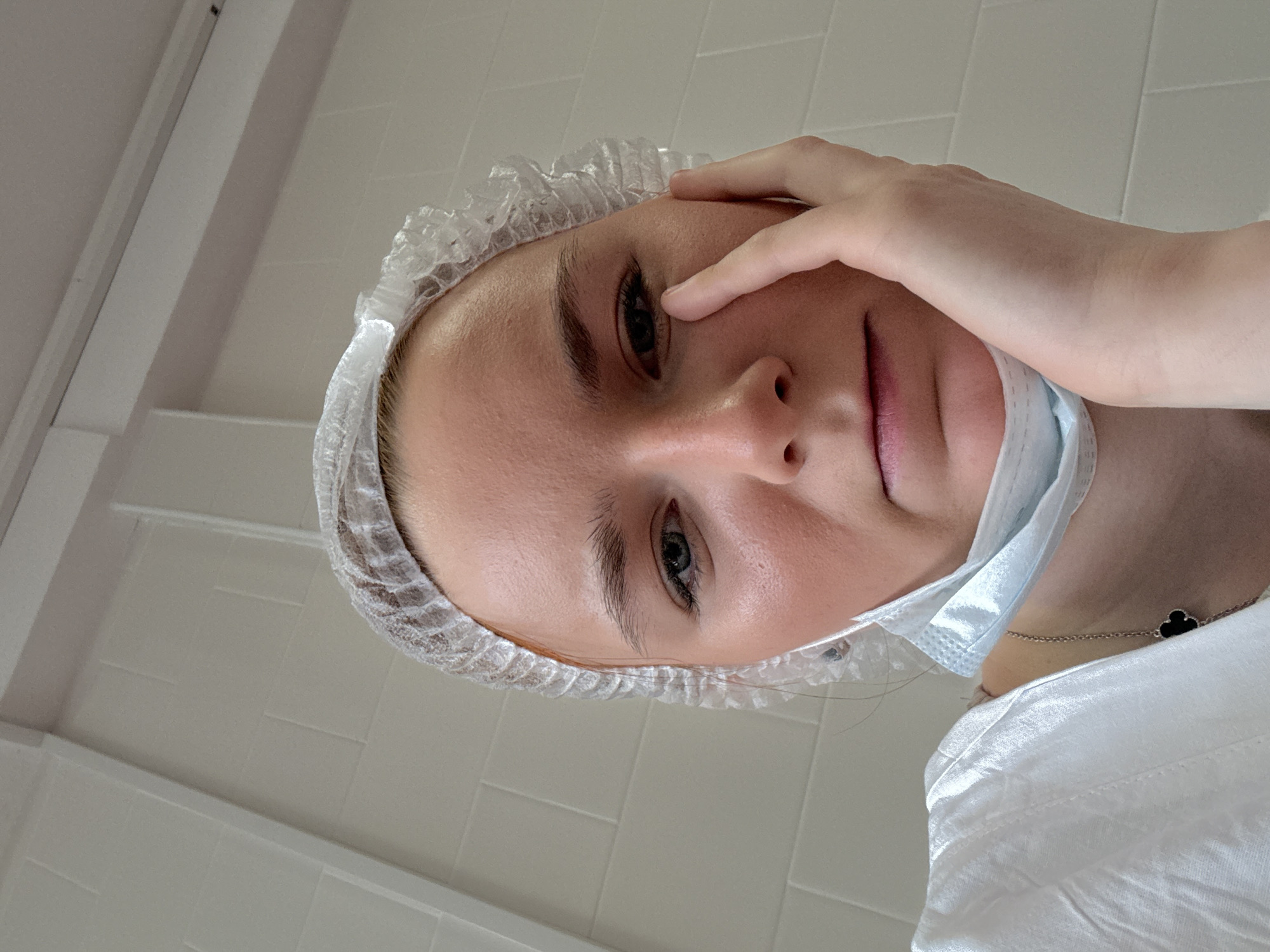 The image shows a woman wearing a disposable hair net and a surgical mask pulled down around her neck. She is touching her face near her eye. The background appears to be a tiled wall, possibly in a clinic or medical setting. A clover-shaped jewelry piece is visible on her neck. The location is Unknown, Russia.