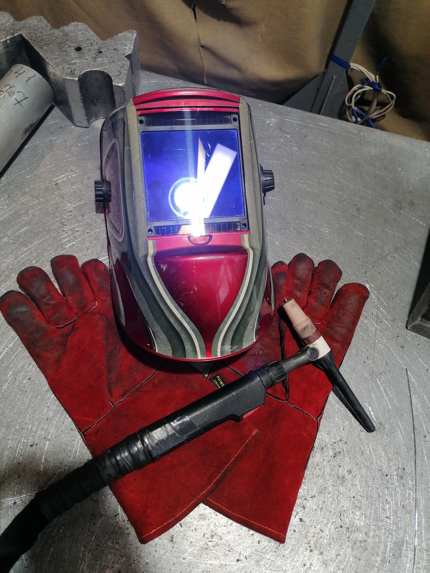 The image depicts a welding setup on a metallic surface. A red welding helmet is positioned on top of a pair of red, well-worn welding gloves. A black welding torch rests across the gloves. Other metallic objects and a tangle of wires are visible in the background.