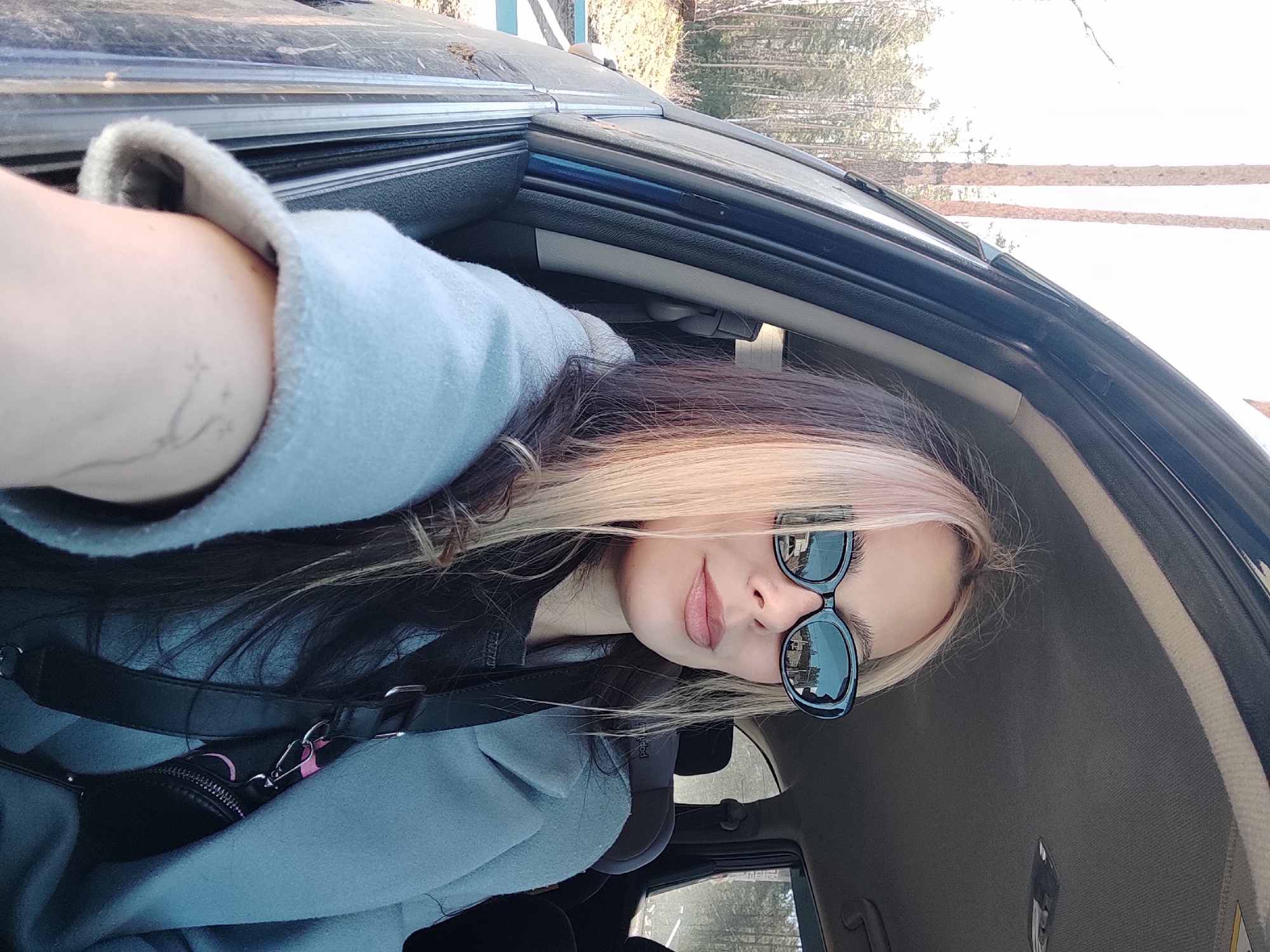 A young woman wearing sunglasses and a light blue coat is taking a selfie inside a car. Her hair is blonde in the front and black in the back. The car is parked near a wooded area. The reflection in her sunglasses shows buildings, which suggests the location is Noviy, Russia.