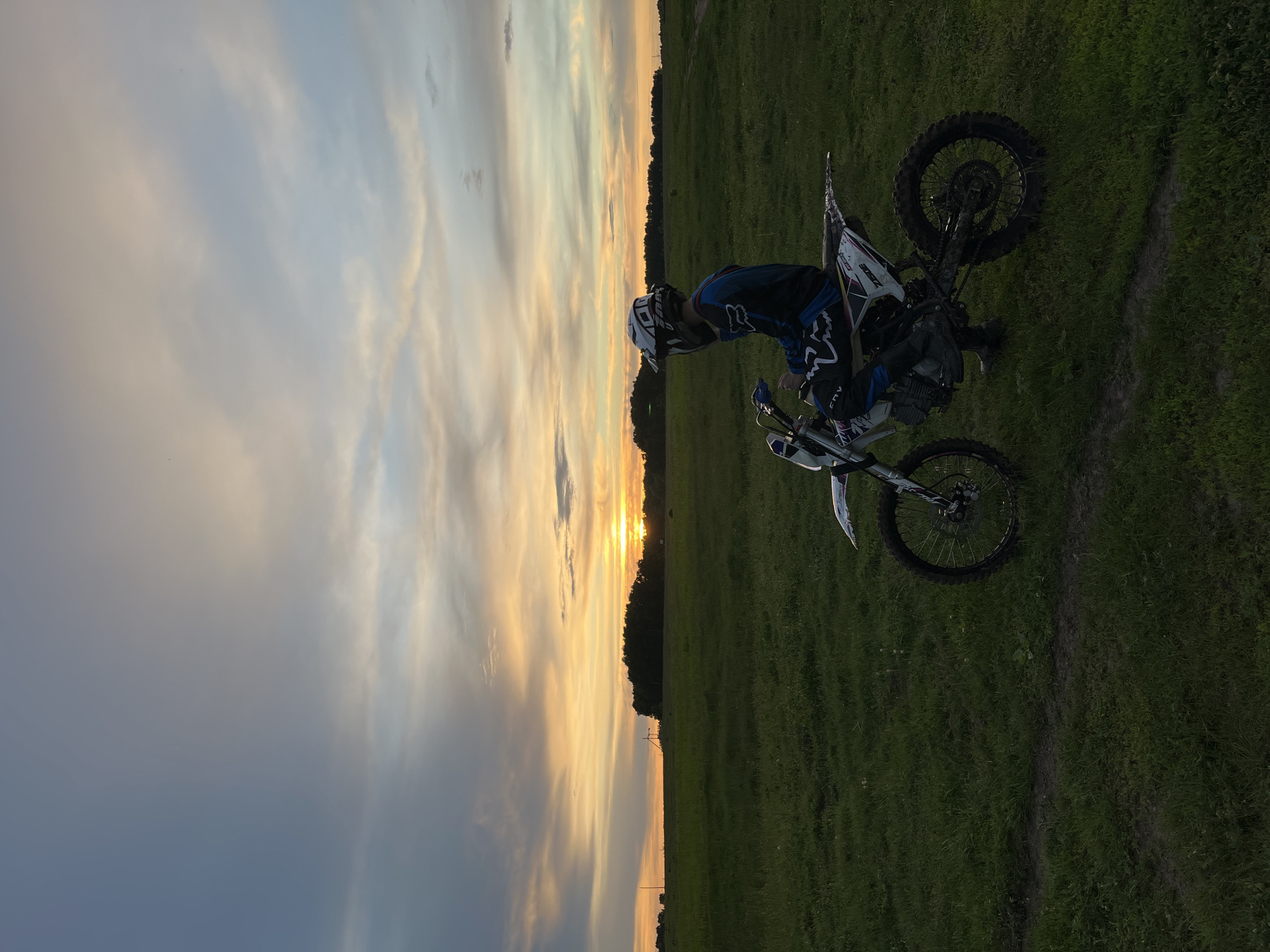 A dirt biker and his motorcycle are positioned on a grassy field. The sunset is visible in the distance, divided by a treeline. The sky displays varying shades of gray, blue, and orange. The biker is wearing a helmet, goggles, and riding gear. The location is near Новочувашский, Russia, indicated by coordinates.