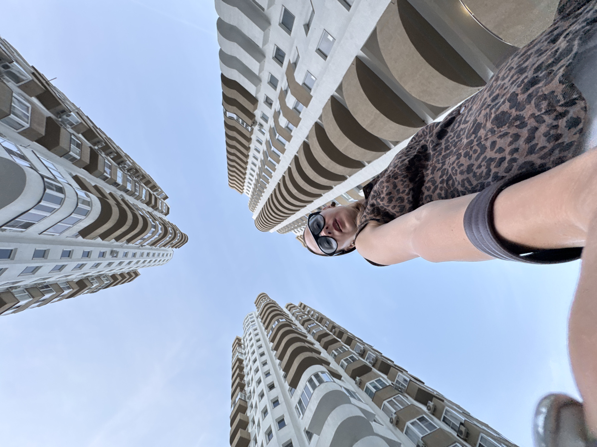 An upward-angle, wide-shot photograph captures a woman in sunglasses, wearing a leopard-print top, looking up at three towering residential buildings in Sochi, Russia. The buildings, characterized by their curved balconies and light-colored facades, dominate the frame against a pale blue sky. The woman is positioned in the lower-right quadrant of the image, with her left arm extending towards the camera, as if taking a selfie. The perspective emphasizes the height and grandeur of the architecture.