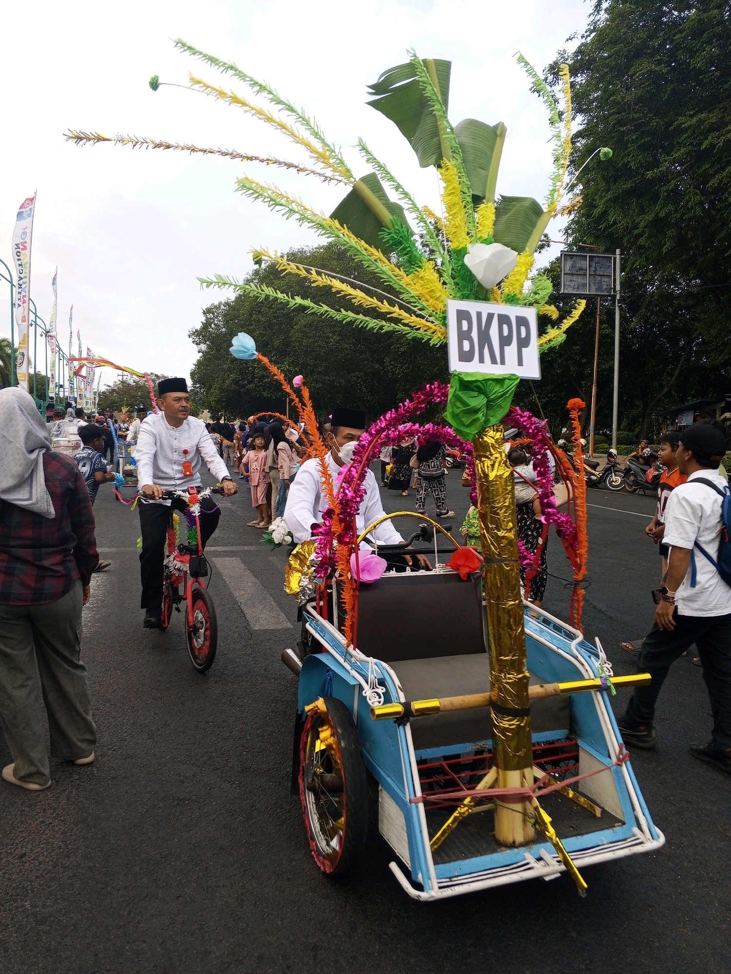 The image depicts a street scene in Banyuwangi, Indonesia, during what appears to be a festive procession. In the foreground, a blue and white pedicab is decorated with colorful streamers and palm fronds, with a sign reading "BKPP" prominently displayed. A person is seated in the pedicab, wearing a white shirt and a black cap. To the left of the pedicab, a man on a decorated red bicycle, also wearing a white shirt and black cap, is pedaling. Several other people, including adults and children, are visible in the background, some wearing traditional Indonesian attire. Flags and banners line the street, suggesting a celebration or public event. The scene is set on a paved road, with trees and buildings visible in the distance. The sky is overcast.