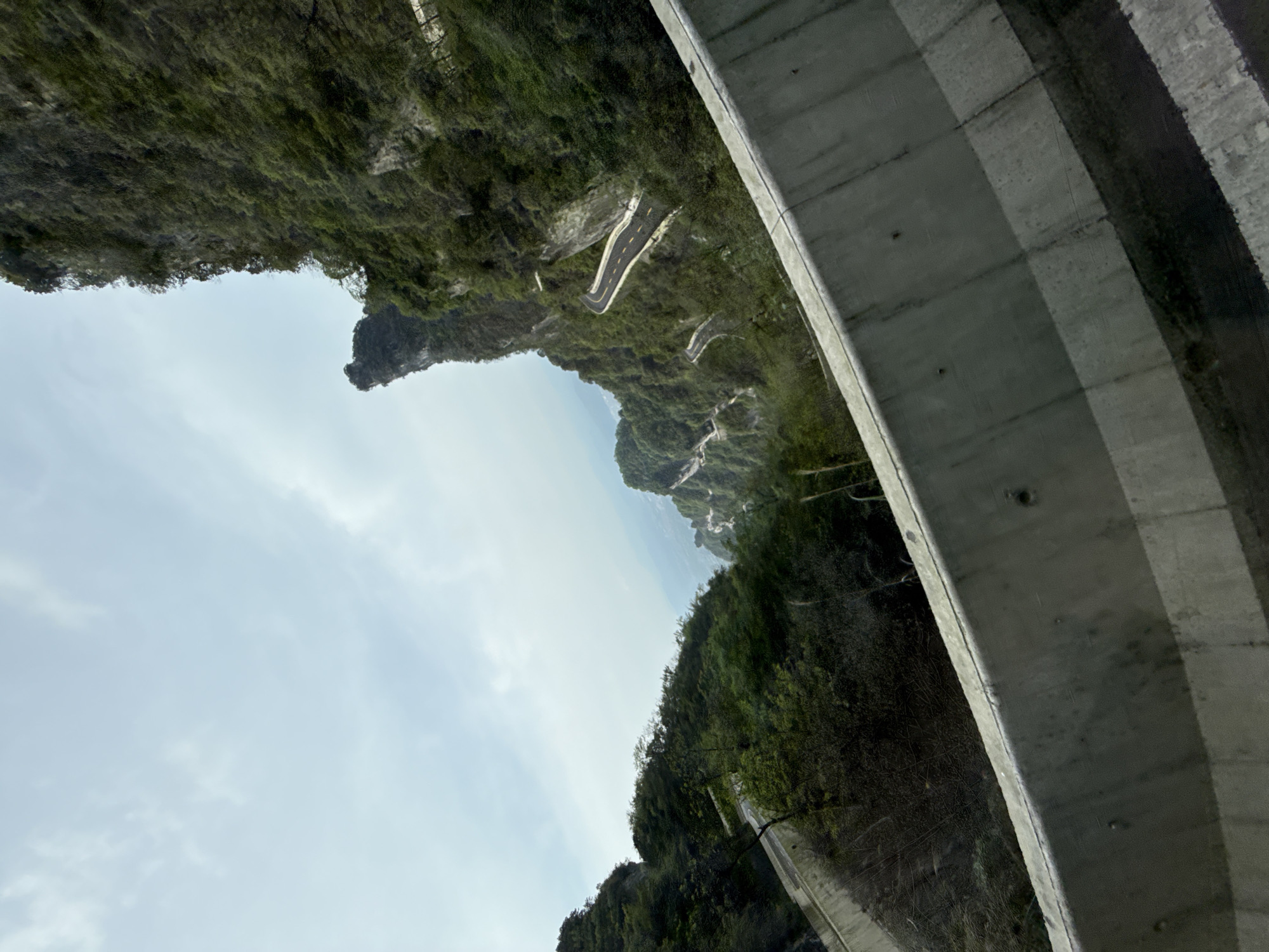 The image shows a view from what appears to be a bridge or elevated structure looking out over a mountainous landscape, likely in the Zhangjiajie area of China. The lower portion of the image is dominated by a grey concrete structure. Beyond this structure, a winding road is visible traversing a lush, green mountainside. The sky is overcast, and the overall composition suggests a high-altitude or mountainous environment with dense vegetation. A prominent rocky outcrop or peak is seen amidst the mountain range.
