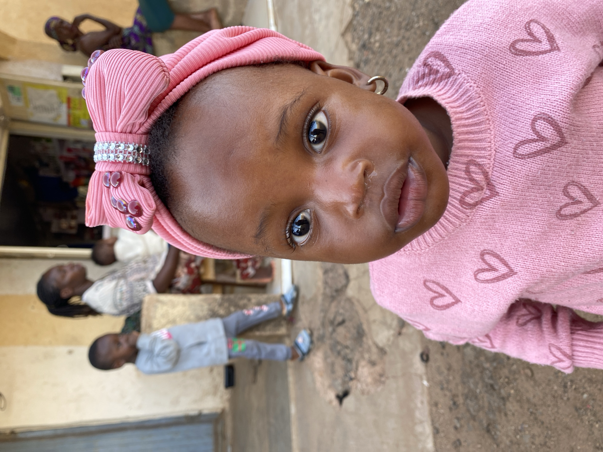 A close-up portrait of a young Black child, likely a girl, is captured in a slightly tilted composition. The child looks directly at the camera with dark, reflective eyes and a calm, curious expression. She is adorned with a prominent pink, ribbed headwrap, fashioned into a large bow at the top, embellished with a sparkling band of silver-colored rhinestones and small decorative beads. A small gold hoop earring is visible in her right ear. She wears a pink knitted sweater patterned with numerous small heart shapes.
The background is softly blurred, revealing an outdoor or semi-outdoor setting characteristic of Vom, Nigeria. Other individuals, both adults and children, are visible in the background, engaged in daily activities. An adult woman in a patterned dress and another child in grey clothing and blue sandals are faintly discernible. The ground beneath them is an earthy, light brown texture. The scene appears to be well-lit by natural daylight, suggesting it is daytime. There is no discernible text visible in the image.