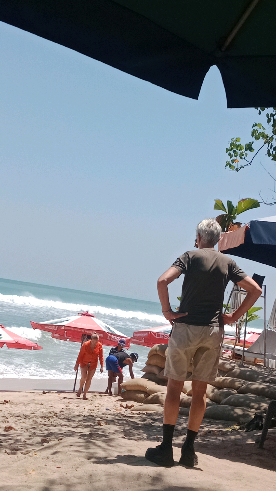 A group of people are on a sandy beach with waves crashing in the ocean. A man with gray hair stands facing away from the camera, hands on his hips. In the background, a woman in an orange shirt walks on the sand, and two men are tending to sandbags. Red umbrellas are set up on the beach. The scene appears to be in Kuta, Indonesia, a popular tourist destination known for its beaches and surf. The sky is clear and blue.