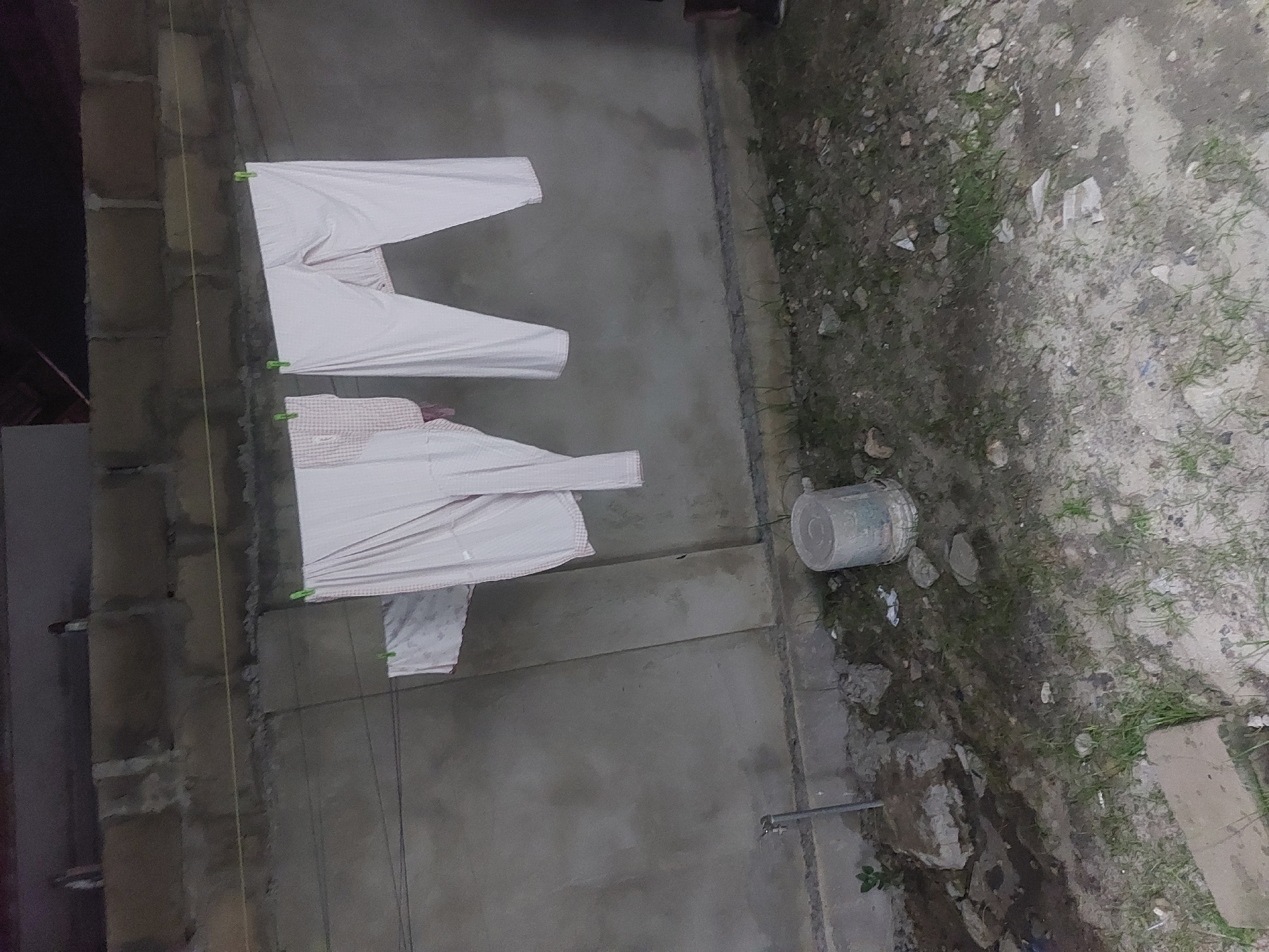 The image shows an outdoor scene, likely in an urban or peri-urban area in Dar es-Salaam, Tanzania, based on the building materials and surrounding vegetation. Clothes are hanging on a clothesline, secured with clothespins, attached to a building with unpainted concrete block walls. A light-colored bucket sits on the ground near the clothesline, and a pipe protrudes from the ground. The ground is a mix of dirt, concrete, and sparse vegetation.