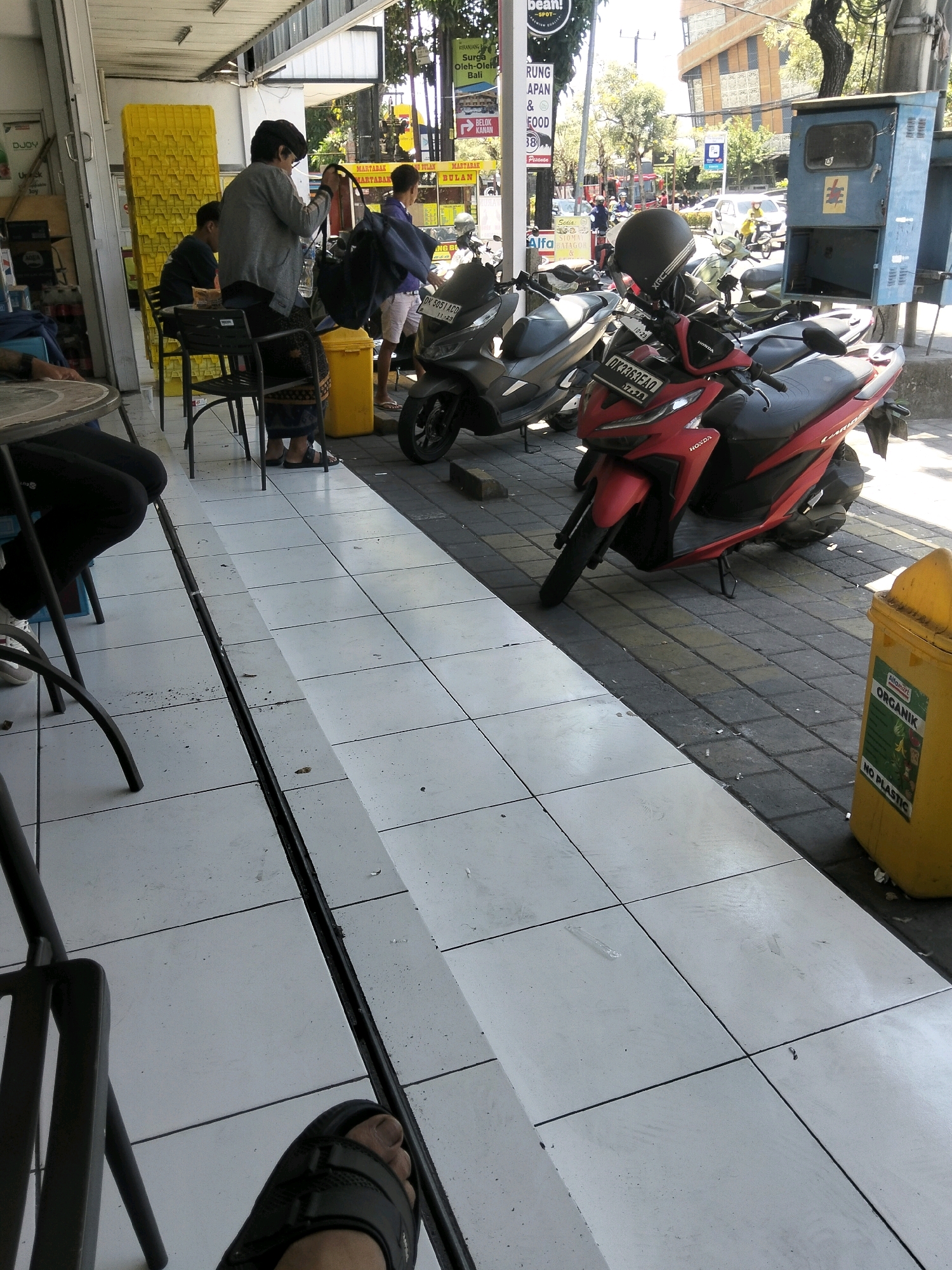 A medium shot captures an outdoor scene in Kuta, Indonesia, focusing on a sidewalk adjacent to a building. The foreground features a person's left foot wearing a black sandal, and a black metal chair leg. The midground displays a red Honda motorcycle with a black helmet on its handlebars, parked next to a grey scooter. A yellow dumpster with "ALFAMART ORGANIK NO PLASTIC" written on it is visible to the right. In the background, several people are standing and walking. A woman in a grey jacket and dark skirt is carrying a black bag, and a young man in a blue shirt stands near her. The building behind them has yellow stacked crates, and signage with Indonesian text is visible. The environment appears to be a busy street with other motorcycles and vehicles in the distance. The ground is paved with light-colored tiles, with a dark strip running down the center of the tiled area. The overall lighting suggests it is daytime, with bright sunlight casting shadows.