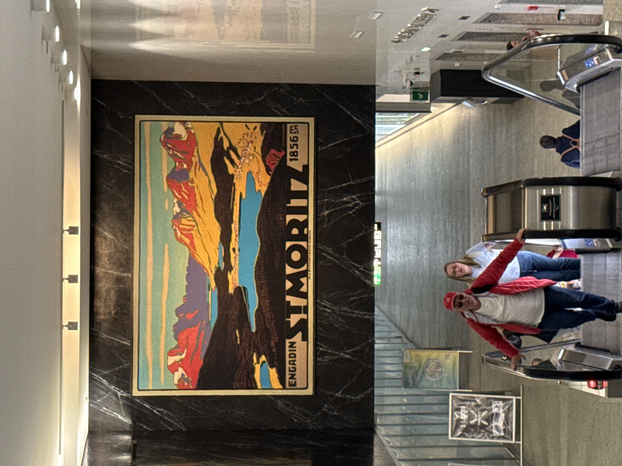 The image is a composite of two photographs. The top image shows a large, framed poster of a landscape with mountains and a lake, set against a dark, marbled wall. Text below the image reads "ENGADIN ST. MORITZ 1856". The bottom image shows an interior scene with people on escalators. In the foreground, a man in a red jacket and baseball cap, and a woman in a blue shirt and jeans are descending on an escalator. Another person, wearing a backpack, is ascending on a separate escalator in the background. Several pieces of artwork are displayed on the walls. The location is identified as the city of Sesto San Giovanni, Italy.