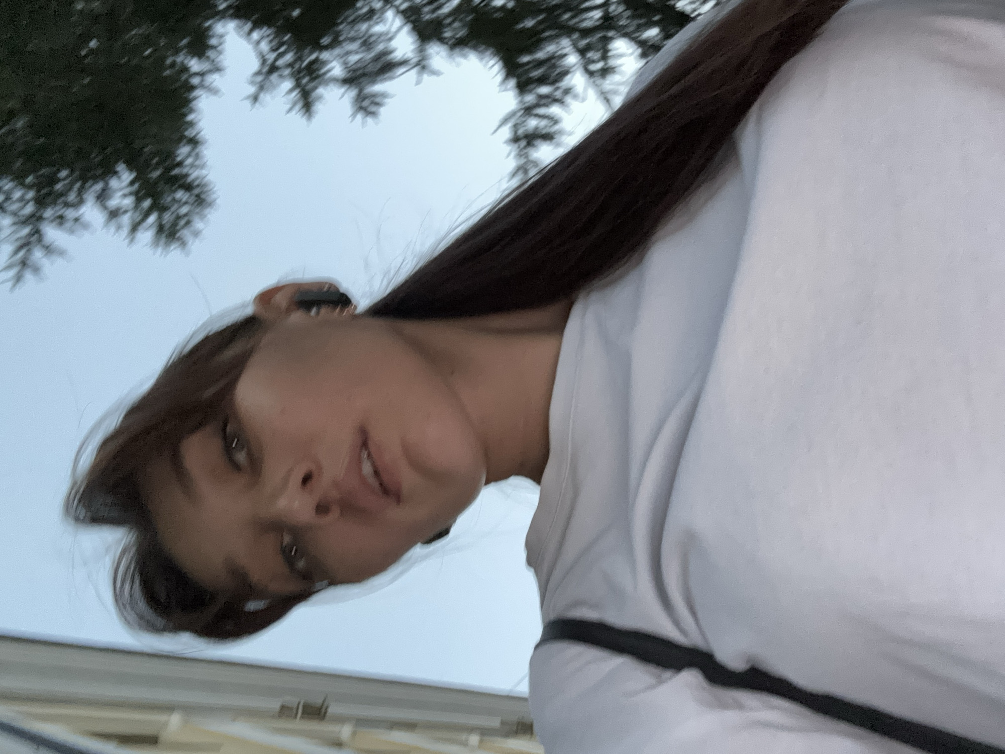 An outdoor, low-angle, close-up selfie shows a young woman from the chest up. She has long, dark brown hair pulled back into a ponytail, and is wearing a white t-shirt. She has earbuds in her ears. The background is a light blue sky with the dark branches of a tree visible on the right side. A blurry building is visible on the left. The image was taken in Novokuznetsk, Russia.