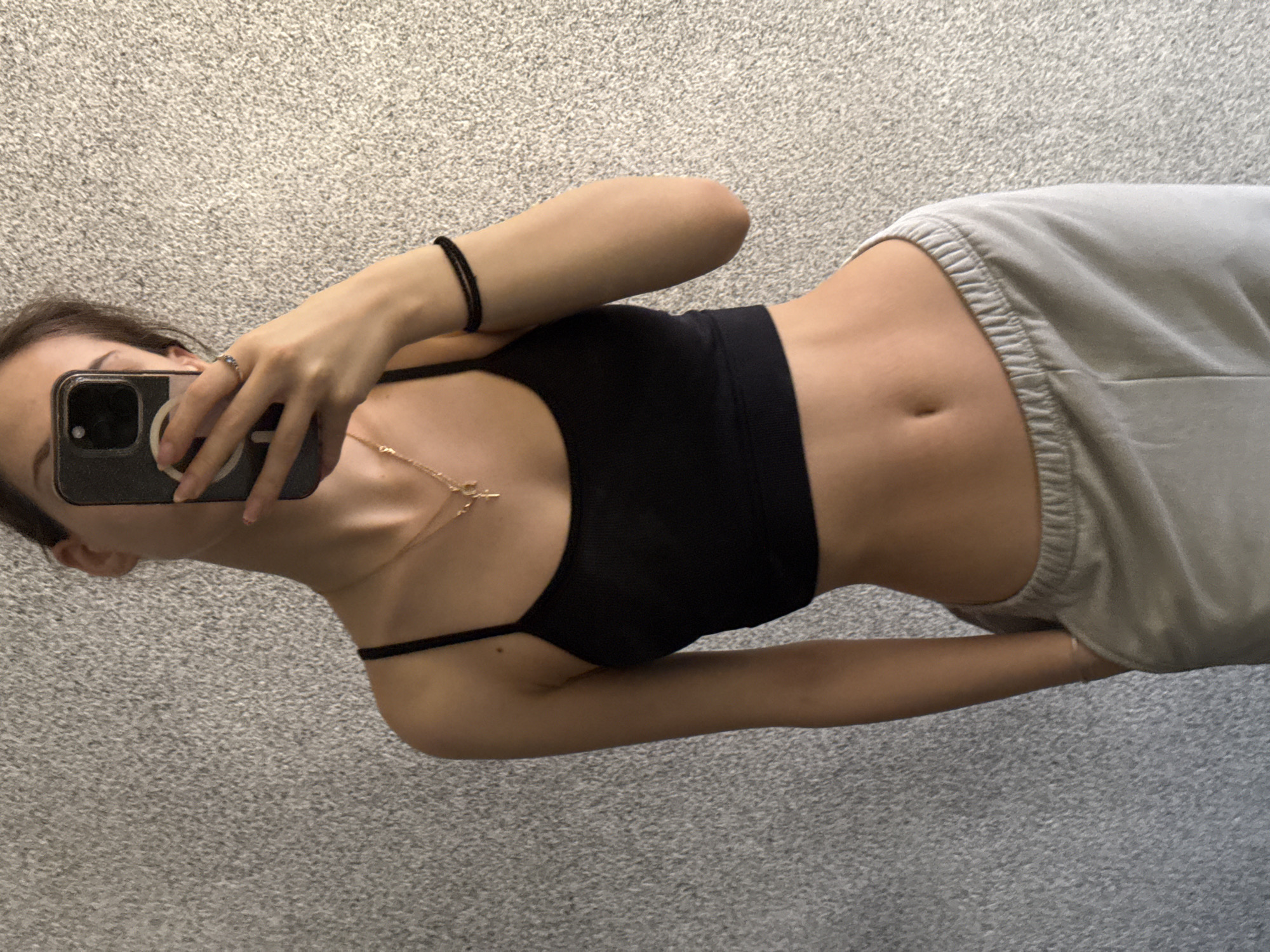 A woman is taking a selfie in an indoor setting. She is wearing a black sports bra and light gray sweatpants, exposing her abdomen. She holds a black smartphone in her right hand, obscuring part of her face. Her left hand is visible, adorned with a ring and a bracelet. A gold necklace is also visible around her neck. The background consists of a gray carpet. There is no information to suggest that this image was taken in Sofia, Bulgaria.