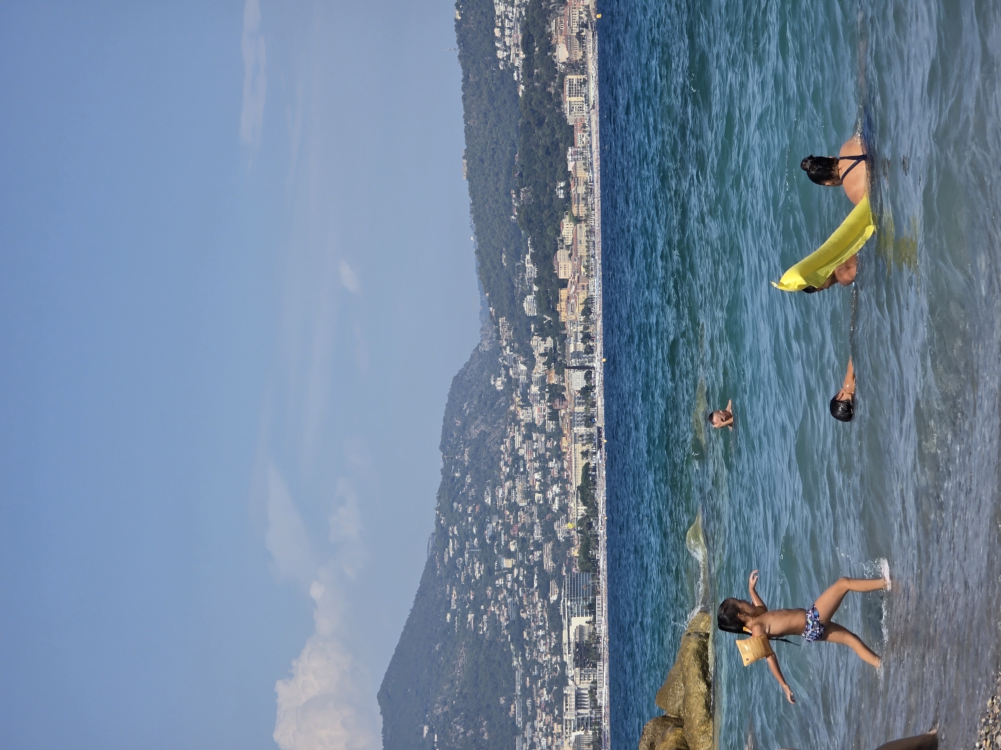 In Nice, France, on a sunny day, several people are enjoying the Mediterranean Sea. A young girl with arm floaties walks in the shallow water near the shore. Further out, two adults are in the water; one is partially submerged with a yellow inflatable raft, and the other is lying on their back. In the background, a densely populated hillside with white buildings and green trees overlooks the coastline, with boats docked in the bay. The sky is a clear blue with a few scattered clouds.