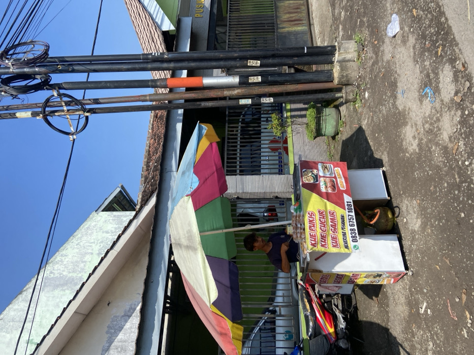 The image depicts a street food vendor in Mojosari, Indonesia, located at coordinates -7.503 latitude and 112.54 longitude. The vendor, a man wearing a black shirt and a cap, is standing behind a white food cart. The cart displays a sign advertising "kue pukis," "kue cucur," and "kue samir." The man is preparing food while standing under a colorful umbrella. The cart is situated next to a building with a white facade and a green gate. In the background, there are four utility poles, one of which has a red stripe. The ground is paved with gray asphalt. There are several items scattered on the ground, including a small tree, a plastic bag, and a blue object. The day is bright, with a clear blue sky.