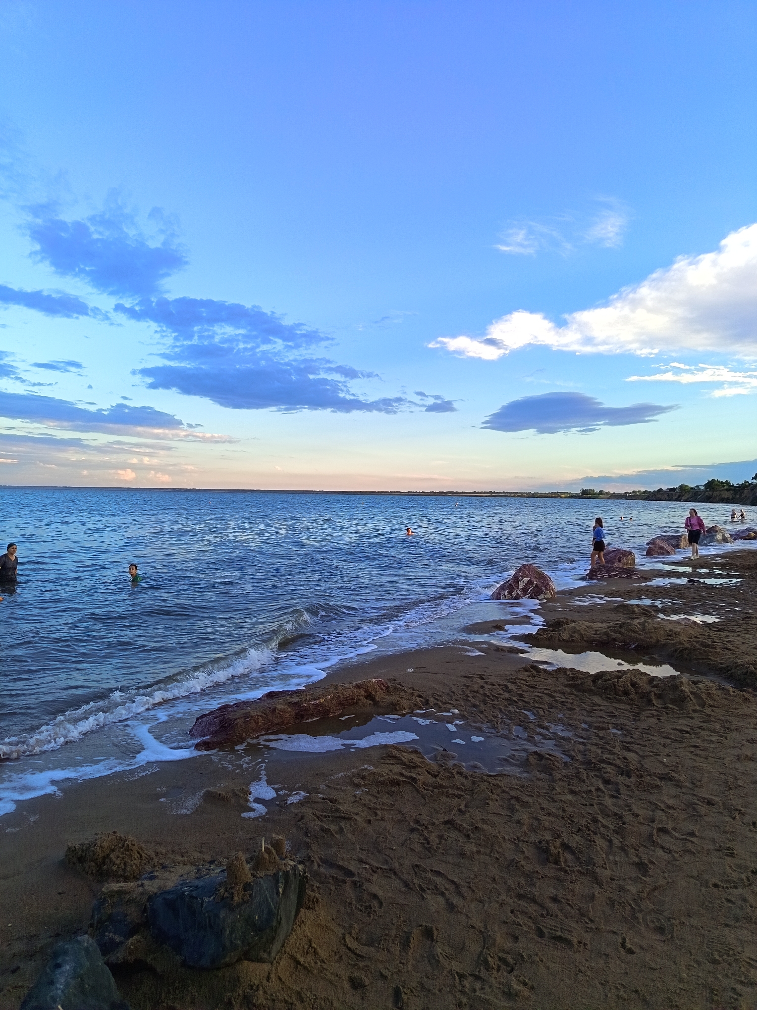 A wide-angle, low-angle shot captures a serene beach scene on a warm day at Lake Kulunda, located in Yarovoye, Russia. The image is divided into three horizontal bands: the sky, the sea, and the shore. The sky, occupying the top third of the frame, is a brilliant blue with scattered white and gray clouds. The sea, filling the middle section, is a deep blue with gentle waves lapping the shore. Several people are visible in the water, some swimming and others standing. The shore, the bottom third, is a mix of dark sand and scattered rocks, with some areas of shallow water reflecting the sky. The lighting suggests it is late afternoon or early evening, with the sun casting a soft glow on the scene. The overall impression is one of peaceful recreation and natural beauty.