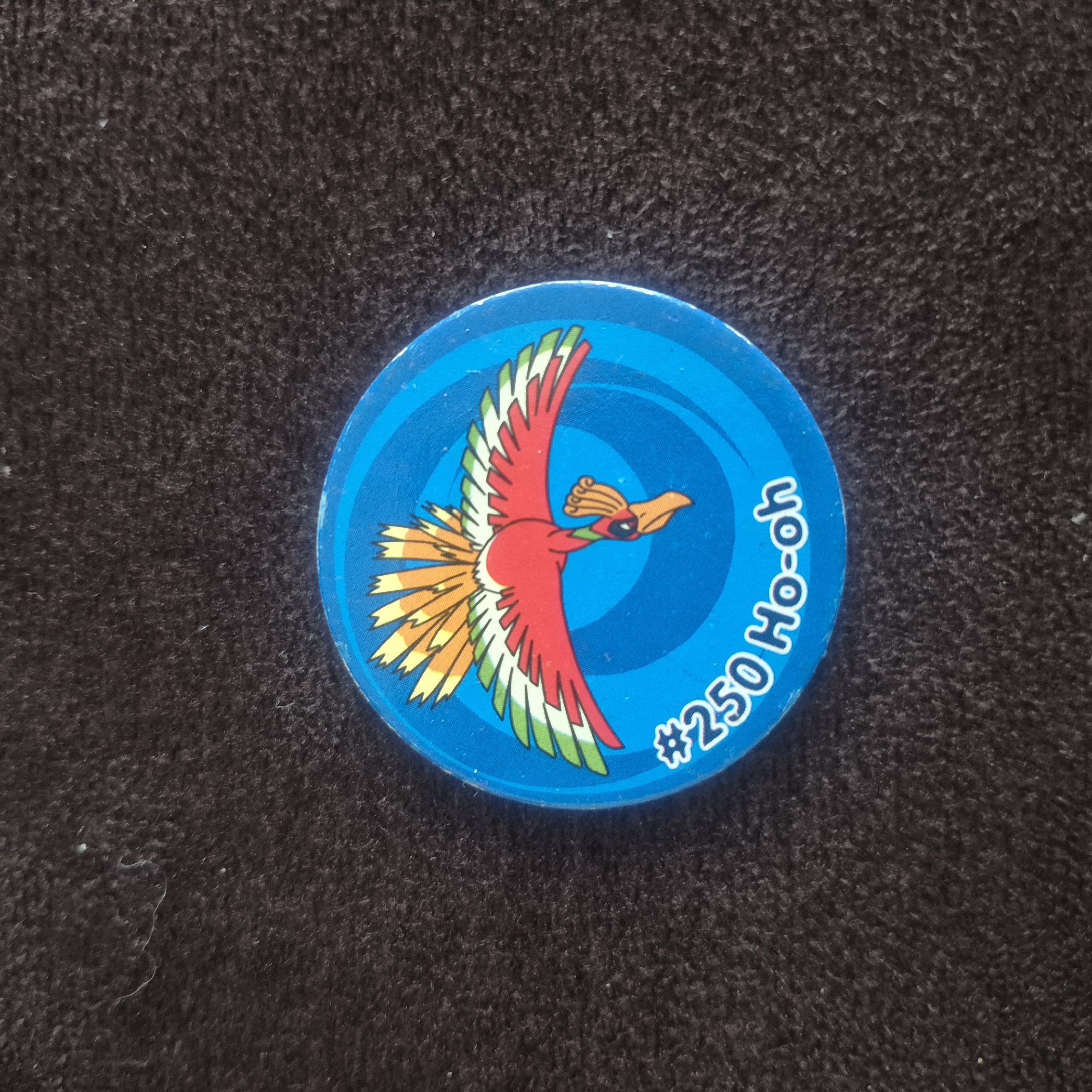A circular blue Pokemon token featuring the character Ho-oh is displayed on a dark, textured surface, possibly a rug or blanket. The token has a white border and depicts the fiery bird Ho-oh in flight against a swirling blue background. The character is rendered in bright colors, with red, yellow, green, and white feathers. At the bottom of the token, the text "#250 Ho-oh" is written in white. The image is taken from a top-down perspective. The setting, Kępno, Poland, is irrelevant to the visual content of the image.