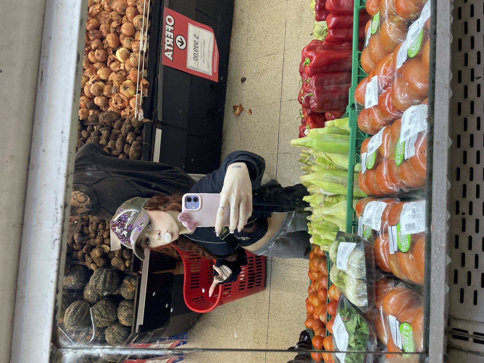 The image is a mirror selfie taken inside a brightly lit grocery store, likely in the produce section, as indicated by the "OFERTA" sign in Spanish, aligning with the provided location context of Buenos Aires, Argentina.
The primary subject is a young person with reddish-brown hair, seen in reflection. They are dressed in a dark hoodie or jacket, grey pants, and a camouflage baseball cap with purple accents. The person has a distinct cheek piercing and dark-painted fingernails. They are holding a smartphone with a pink case and a small pink accessory, using it to capture their reflection. A red shopping basket is visible in their hand, suggesting they are in the process of grocery shopping.
The setting is a supermarket produce aisle, filled with various fresh goods. To the left of the person's reflection are large piles of onions and potatoes, with darker, squash-like vegetables below. The shelving unit being used as a mirror reflects rows of red bell peppers, bundles of corn on the cob, and numerous packaged items. These include tomatoes in clear plastic, some with labels showing codes like "GK10," "25.180," and "2023," along with packaged greens. Eggplants are partially visible at the bottom.
A red sign, reflected as "ATЯЗНО," clearly reads "OFERTA" (Offer) and displays a price of "2.999,00," likely in Argentine Pesos. Below the price, text that appears to be "CEBOLLA Y PIMIENTOS X KG" (Onions and Peppers per Kg) is visible, though slightly distorted by reflection. The word "BREATHE" can be seen on the sole of the person's reflected shoe. The overall scene depicts a casual shopping trip within a well-stocked supermarket.