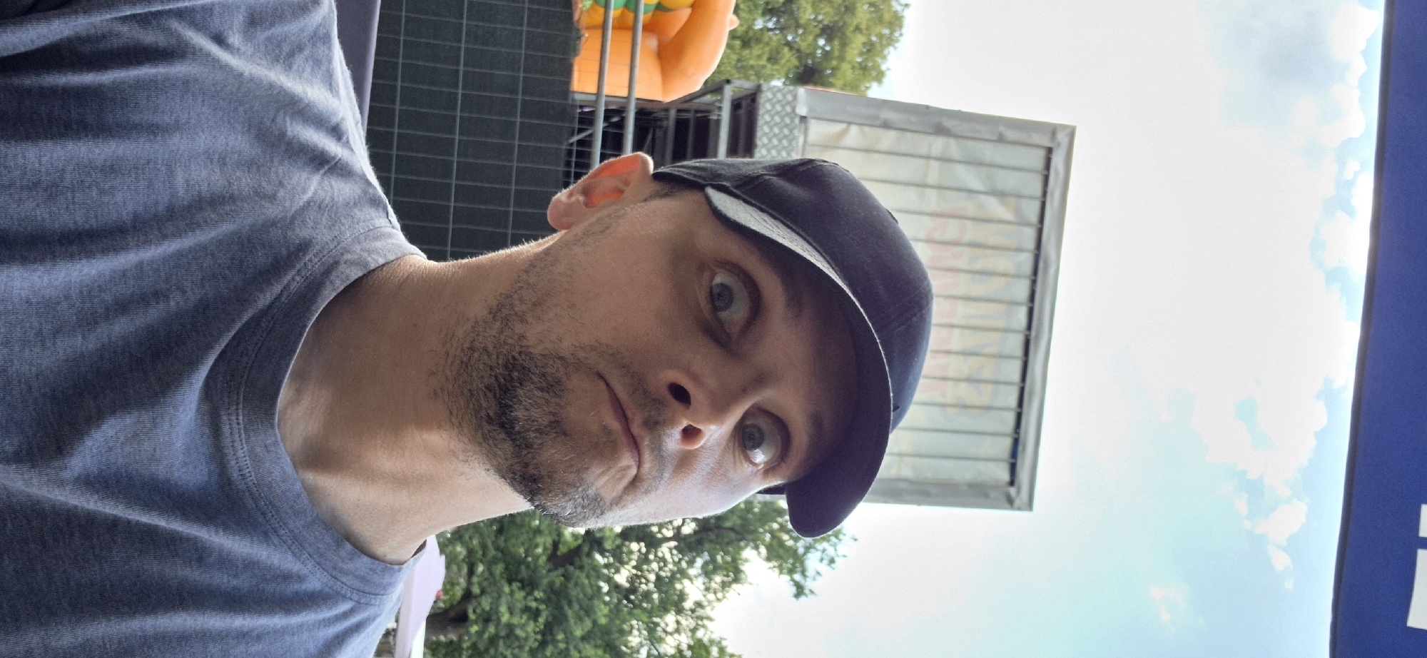 A close-up, selfie-style shot captures a man wearing a gray t-shirt and a black baseball cap. He has light-colored eyes and a short beard, and his facial expression is one of mild surprise or apprehension, with his mouth slightly open and eyebrows raised. His head is tilted back, and the camera is positioned above him, looking down.
In the background, an inflatable orange structure, possibly part of an amusement or play area, is visible on the left. Behind that, a dark fence with a diamond pattern runs horizontally. Further in the background, a large, silver, rectangular structure, possibly a trailer or stage, is mounted on a platform. Trees with green foliage frame the scene, and a bright sky with scattered clouds is visible above. A dark blue banner or awning runs across the very top of the image. The overall setting appears to be an outdoor event or festival. The man's clothing and the casual atmosphere suggest he is attending a public gathering. The image was taken during daylight hours. The location is identified as Gdańsk, Poland.