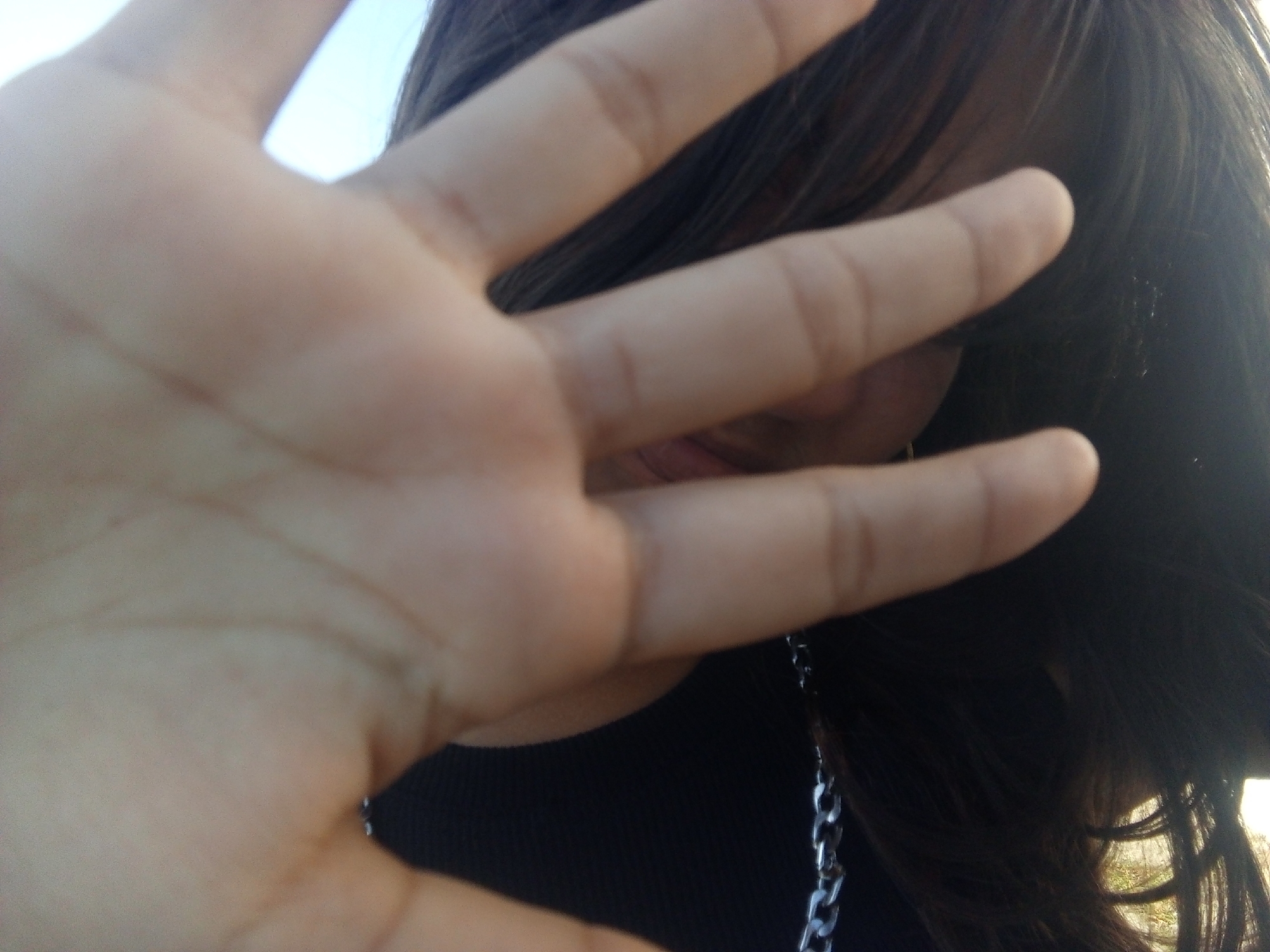 A photograph taken outdoors, likely in daylight, features a hand held up to obscure a person's face. The hand is fair-skinned, with visible finger joints and lines on the palm. The person behind the hand has dark hair and is wearing a black top and a silver chain necklace. The mouth is slightly visible between the fingers. The location is presumed to be Juan Griego, Venezuela. The background is out of focus. The composition suggests the person with the hand is intentionally blocking the camera's view.