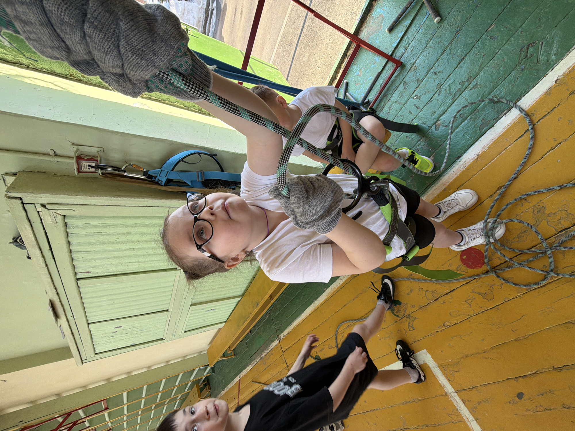 The image shows an indoor climbing activity, likely in a school or gymnasium in Izhevsk, Russia. A young person wearing glasses, a white t-shirt, black shorts, and climbing gear is holding onto a rope and appears to be suspended or climbing a wall that is visible behind them. Another child, also in climbing gear, is partially visible on the climbing wall in the background. Another child in black shorts and a black t-shirt is standing on the floor. The floor is wood and painted in yellow and green. A gray rope is on the floor near them. The wall is light green.