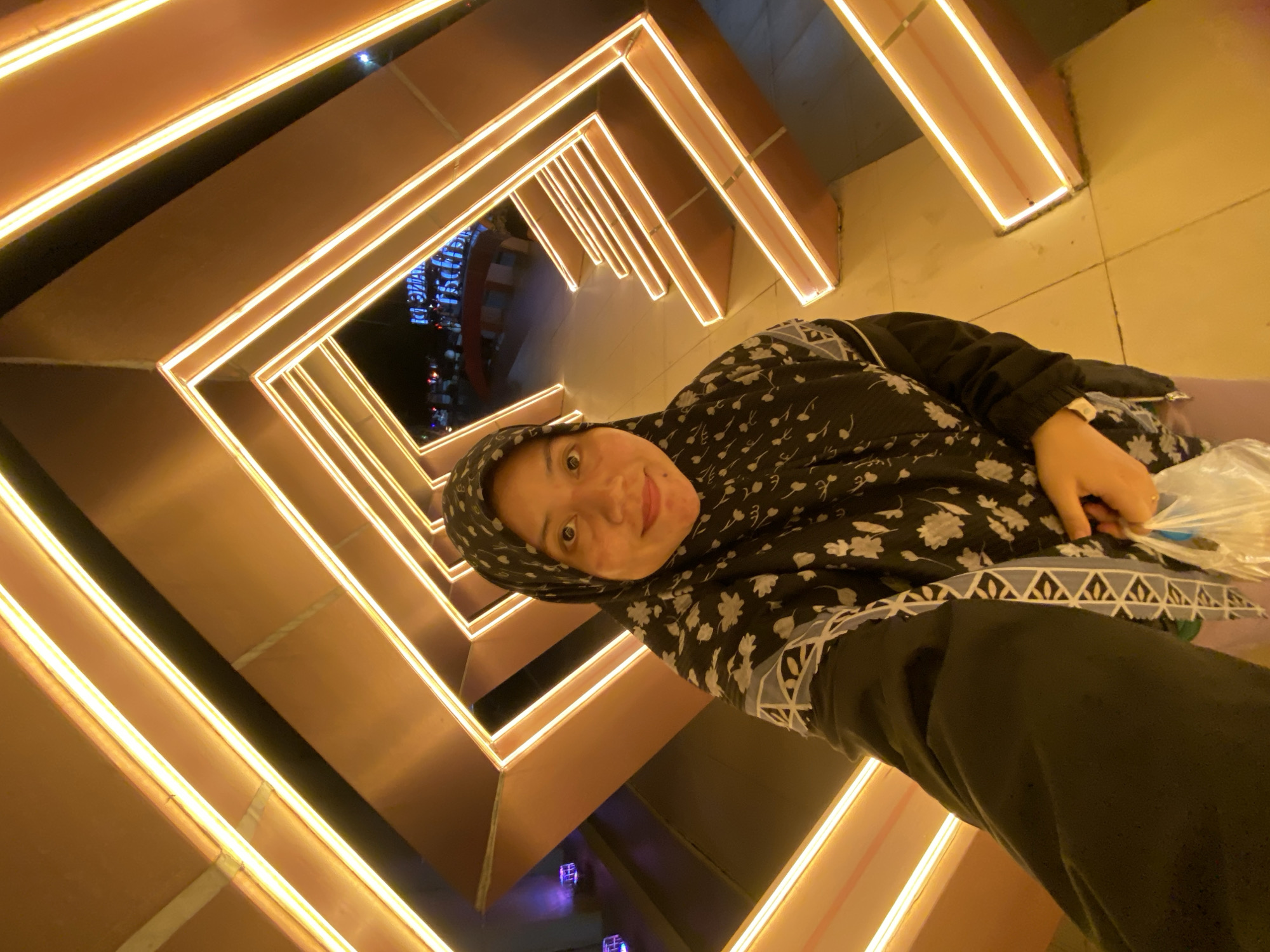 In Banjarbaru, Indonesia, a person wearing a black hijab with a floral pattern takes a selfie in an illuminated pathway. The pathway is constructed with angular, metallic structures outlined by bright yellow lights. The person is holding a clear plastic bag in their right hand. The ground appears to be tiled. In the background, distant blue lights and a street scene are visible.