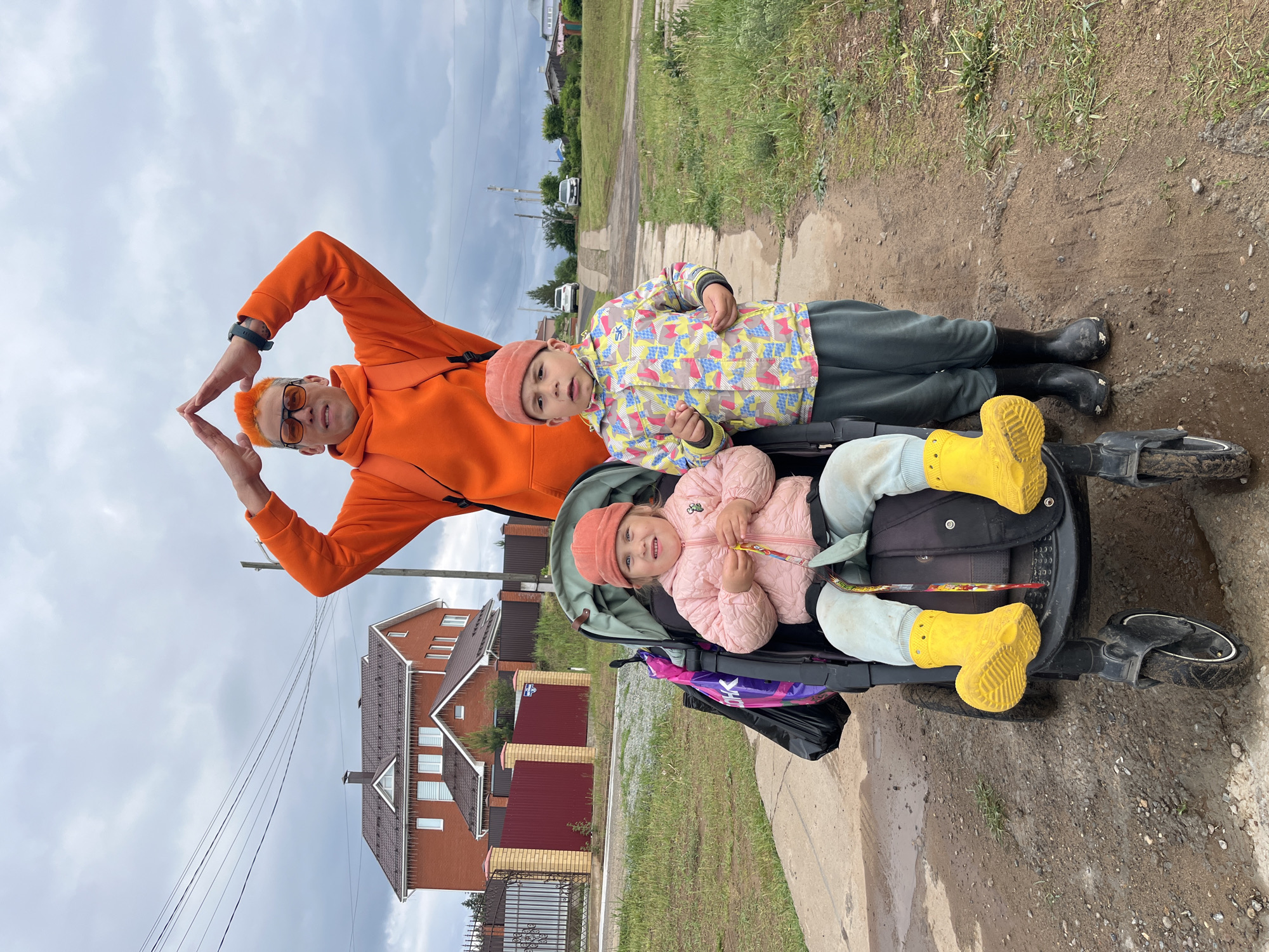 An outdoor shot shows a man and two children in Chaikovsky, Russia, on a cloudy day. The man, wearing an orange hoodie and sunglasses, forms a triangle with his hands above his head. A child in a multicolored jacket and black boots lies on top of a stroller, while another child in a pink jacket and yellow boots sits inside it. The background includes a house with a red roof, a grassy area, and a sidewalk. The ground near the stroller appears muddy.